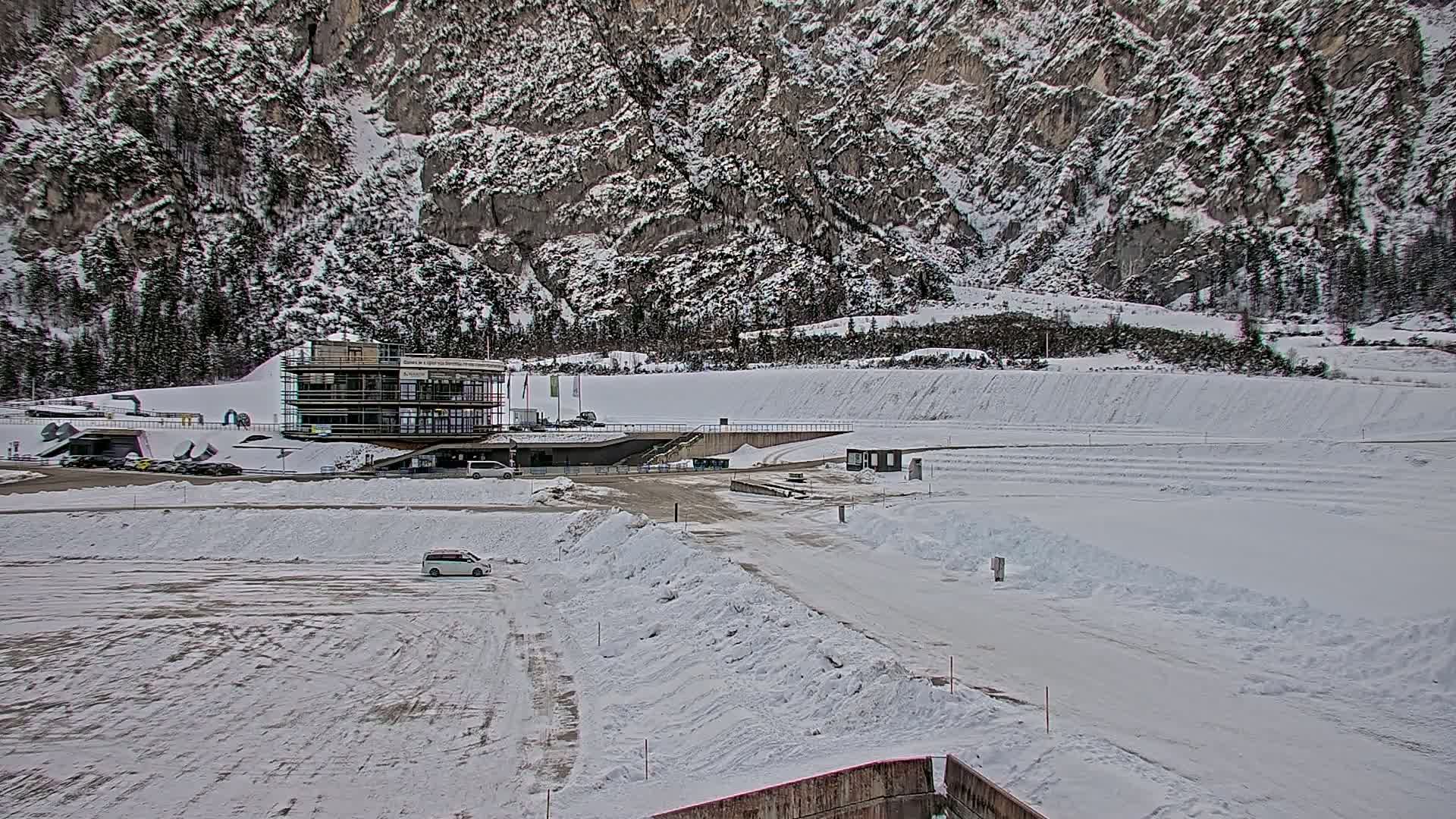 A snow-covered parking lot and roadway are situated in front of a building under construction, with a large snow-covered mountain rising behind it.