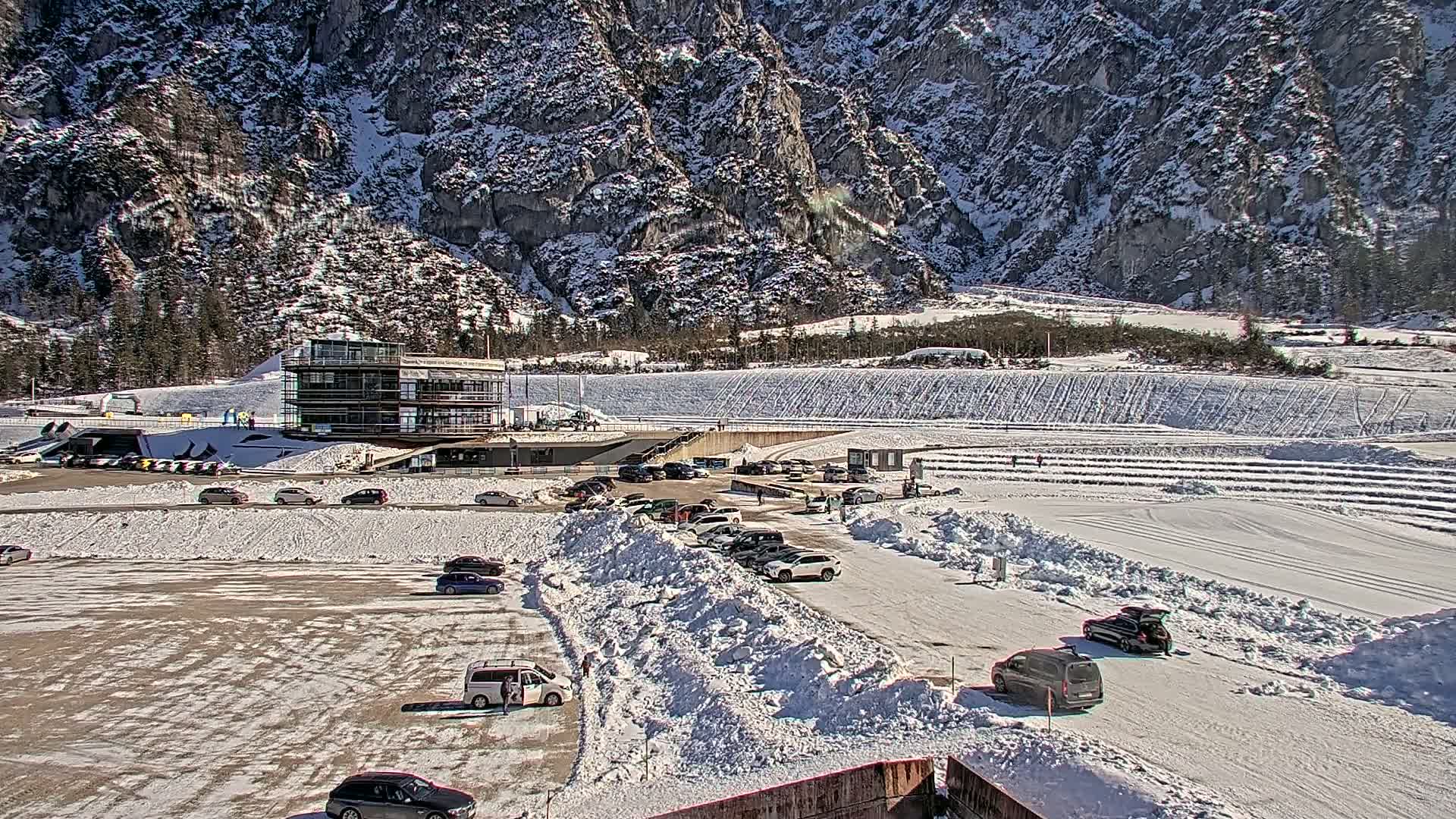 A snowy parking lot with cars, next to a modern building, sits at the base of a large, snow-covered mountain range on a sunny day.