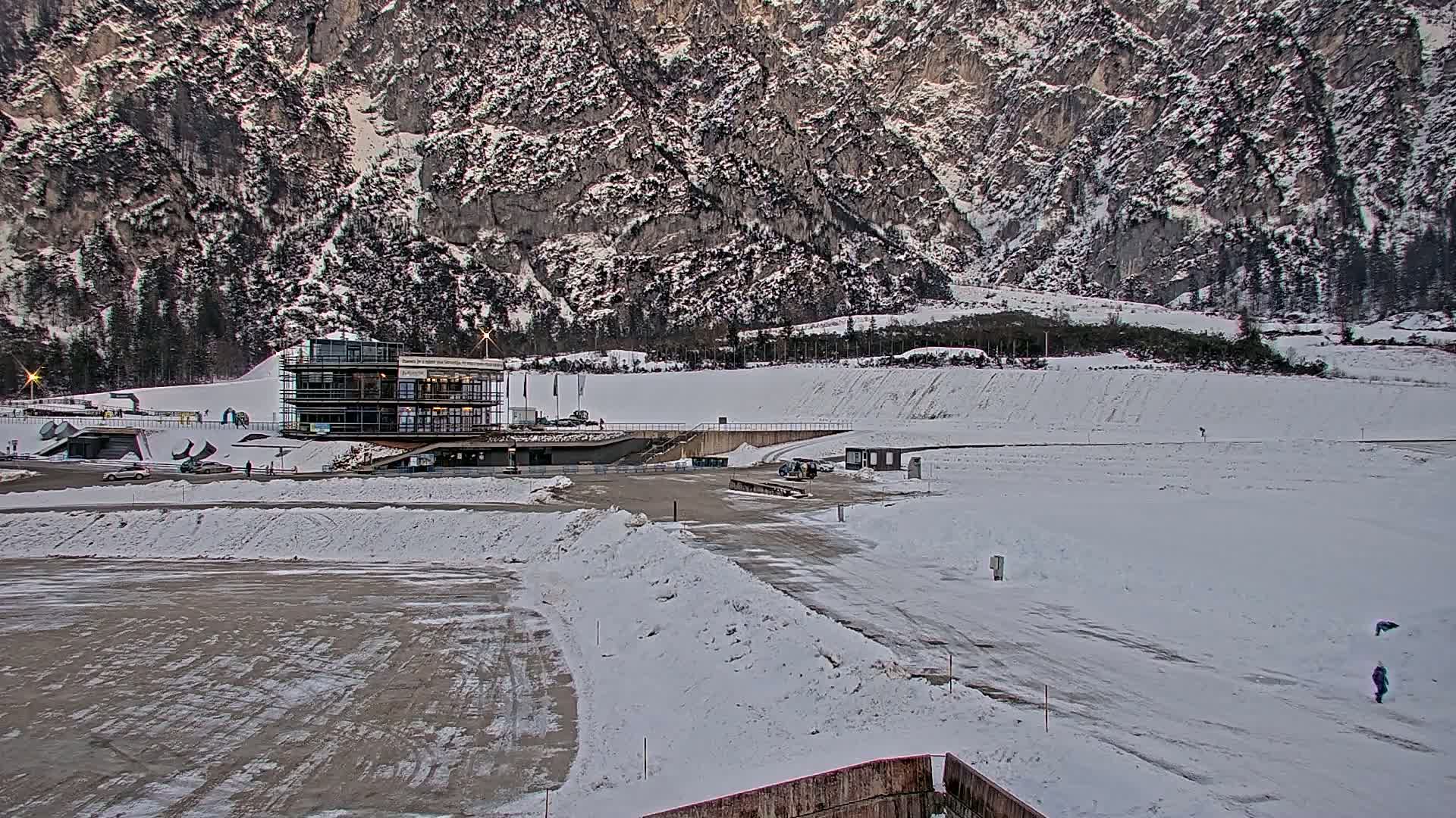 A snowy landscape features a modern building near a large, snow-covered field, all set against a backdrop of a snow-dusted mountain range.