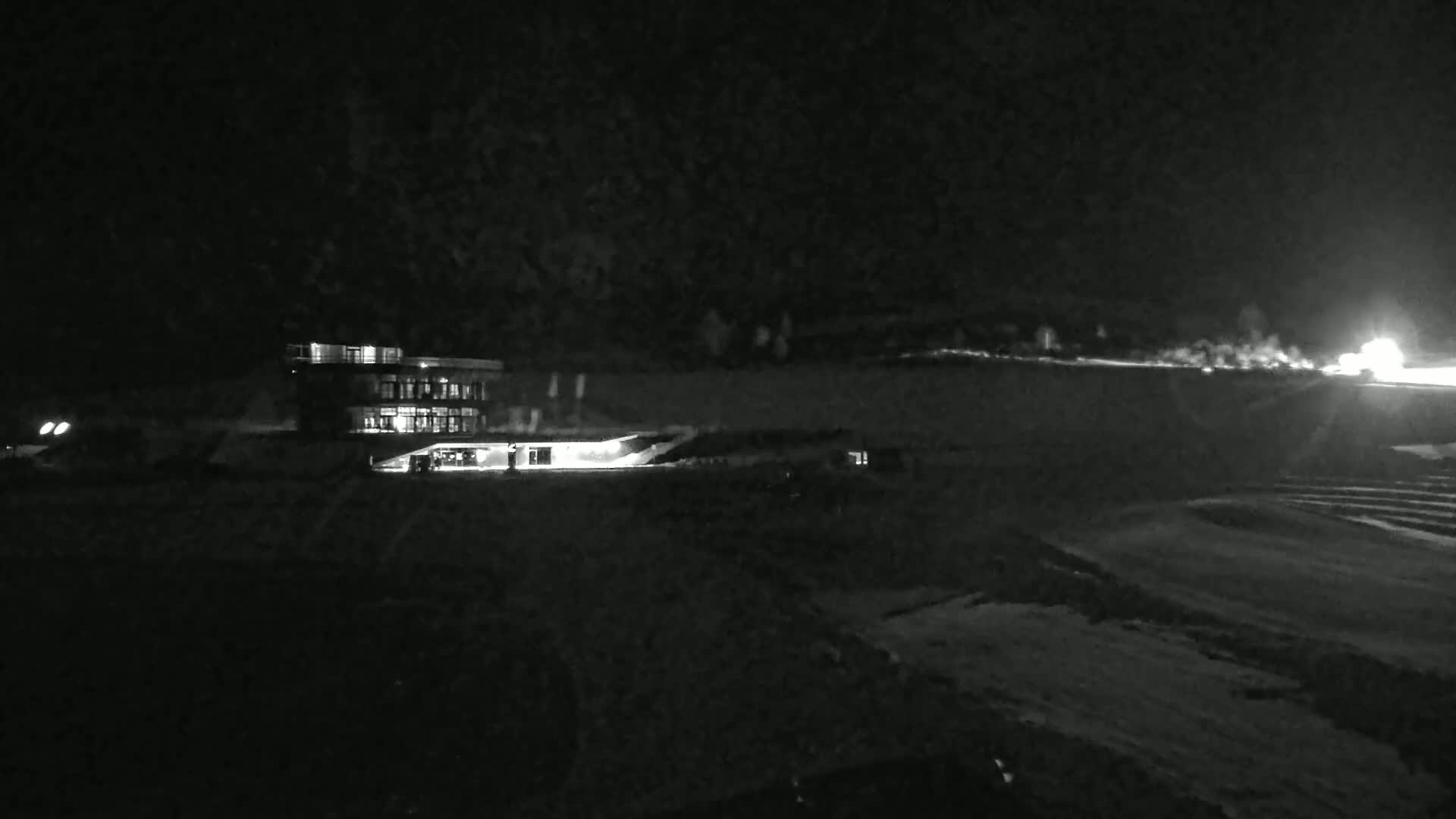 A brightly lit building stands in a dark, snowy landscape at night, with a distant light source visible in the background.