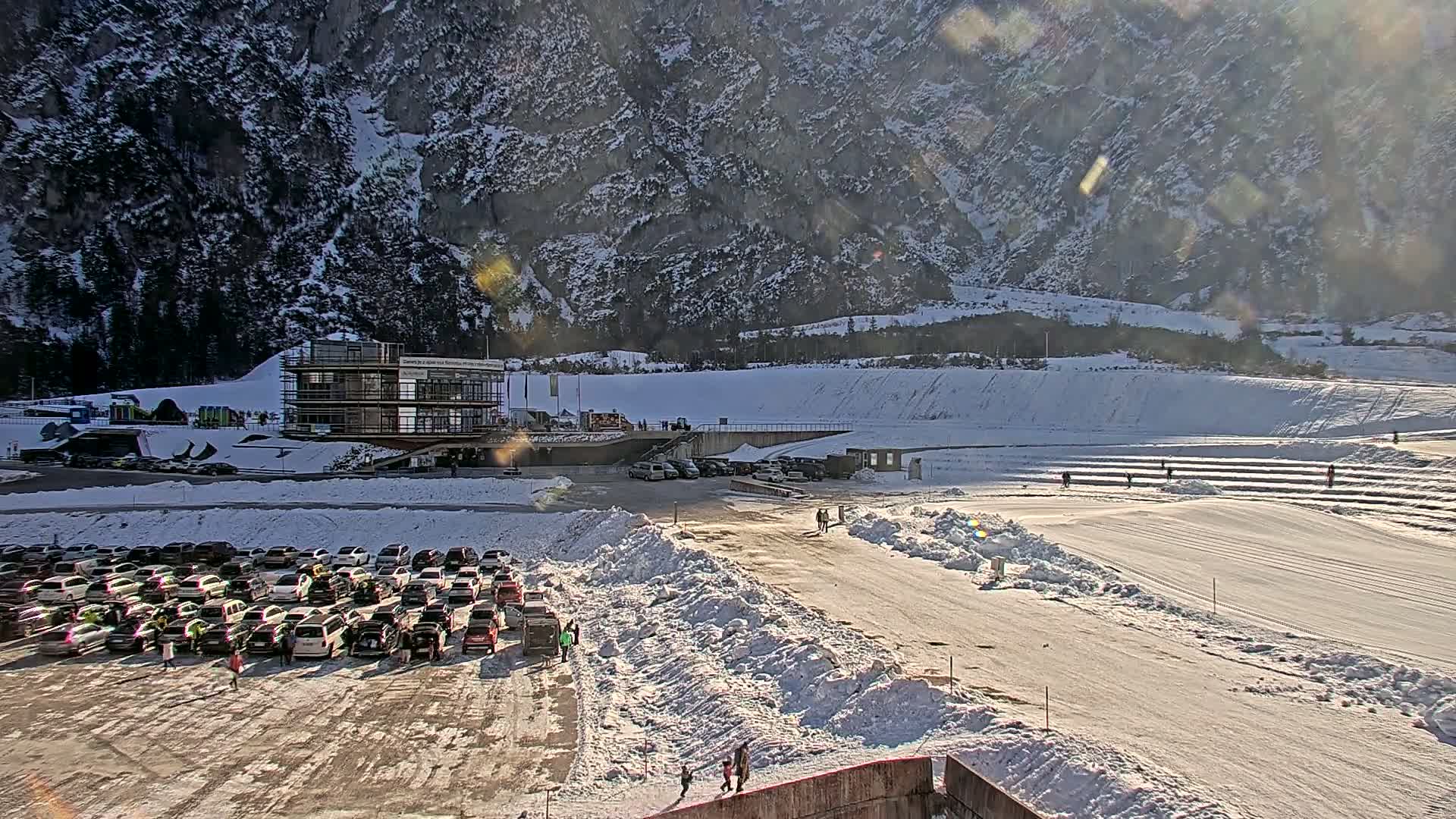 A large parking lot filled with cars sits in front of a partially-constructed building, next to a snow-covered track or course, all nestled at the base of a large, snow-dusted mountain under sunny conditions.