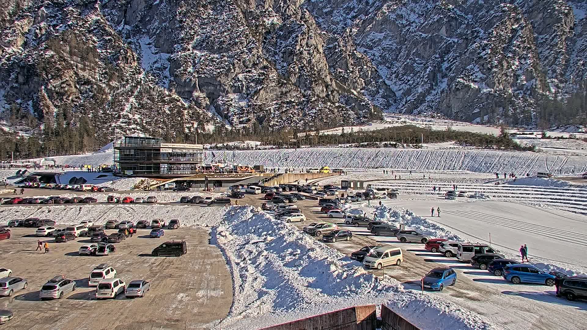 A large parking lot filled with cars sits below a multi-story building next to a snowy, mountainous landscape on a sunny day.