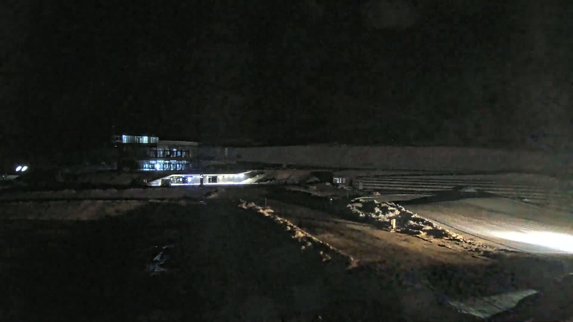 At night, a brightly lit building and some machinery are visible in a mostly dark, snow-dusted construction area.