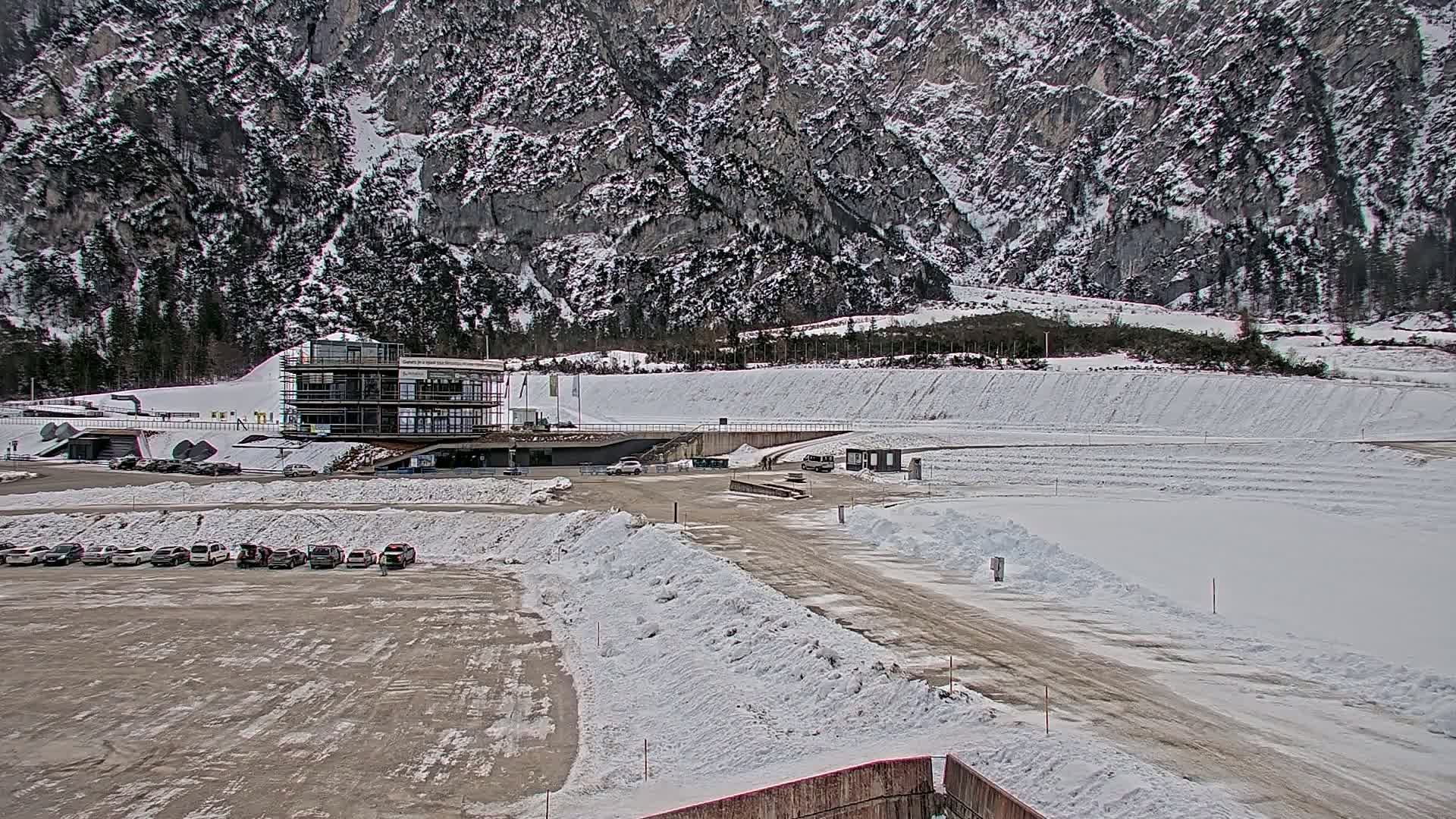 A snow-covered parking lot with several cars sits before a modern building at the base of a large, snow-dusted mountain.