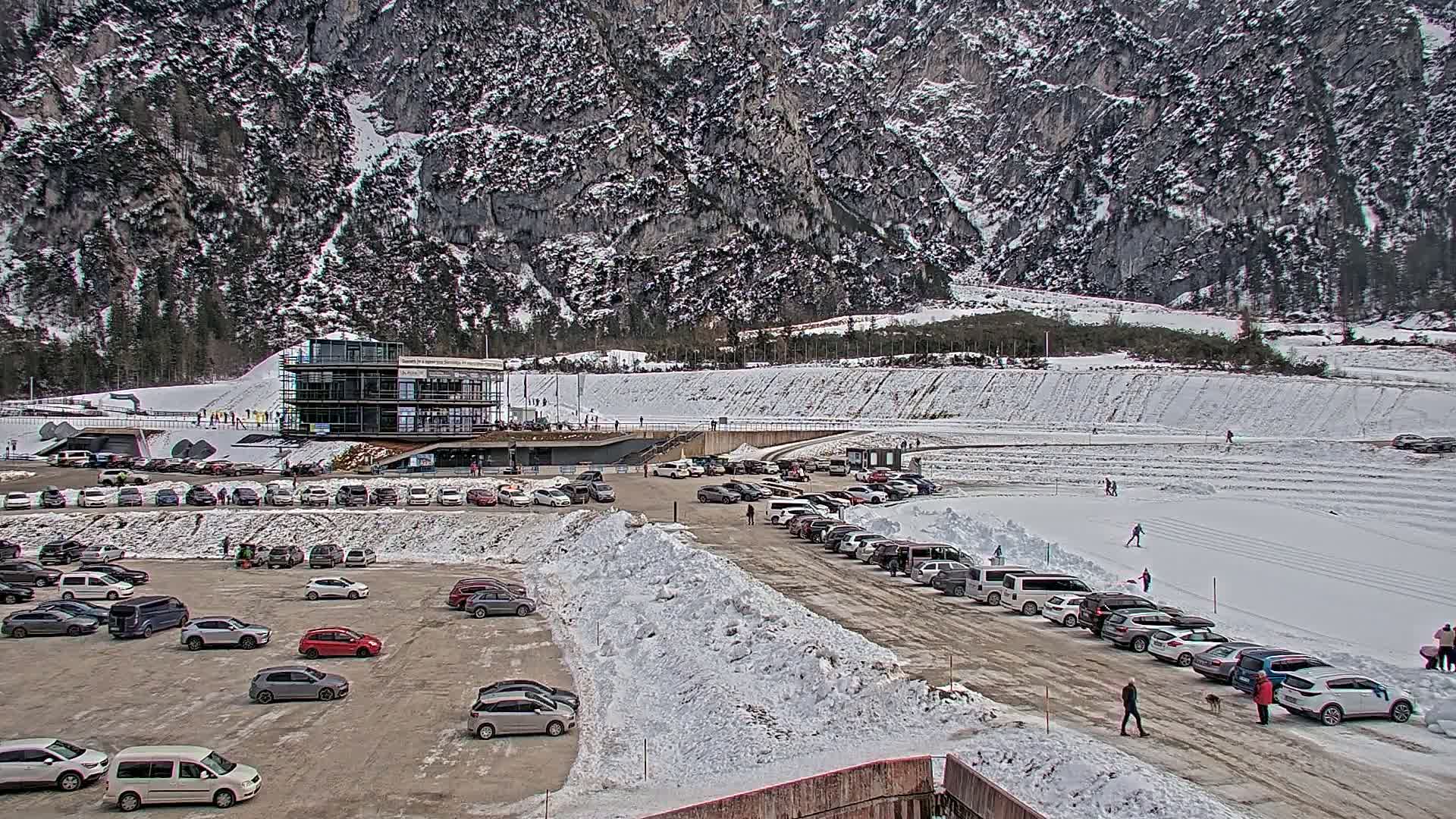 A large parking lot filled with cars is situated below a modern building, which is set against a snowy mountainside with people cross-country skiing in the background.