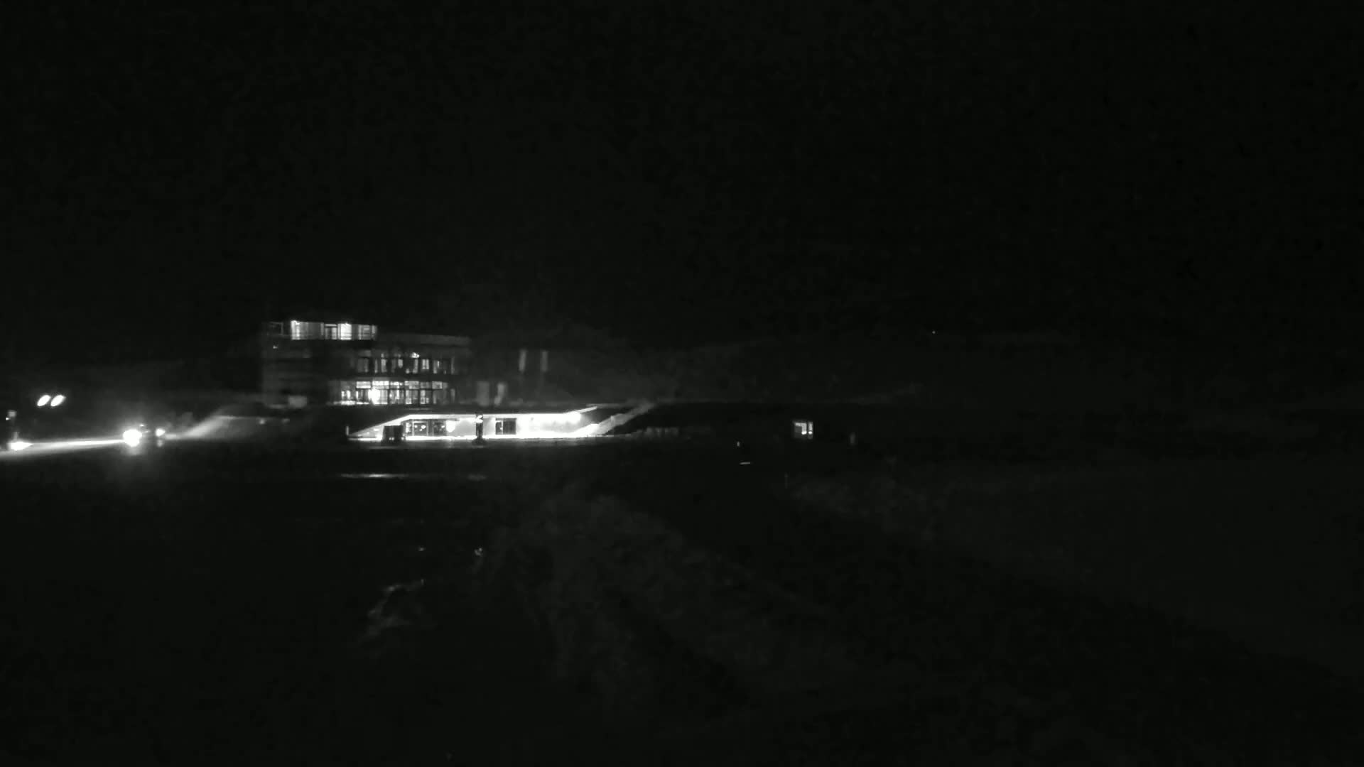 A brightly lit building is visible at night, with a couple of vehicles in the foreground, possibly on a road, in snowy conditions.