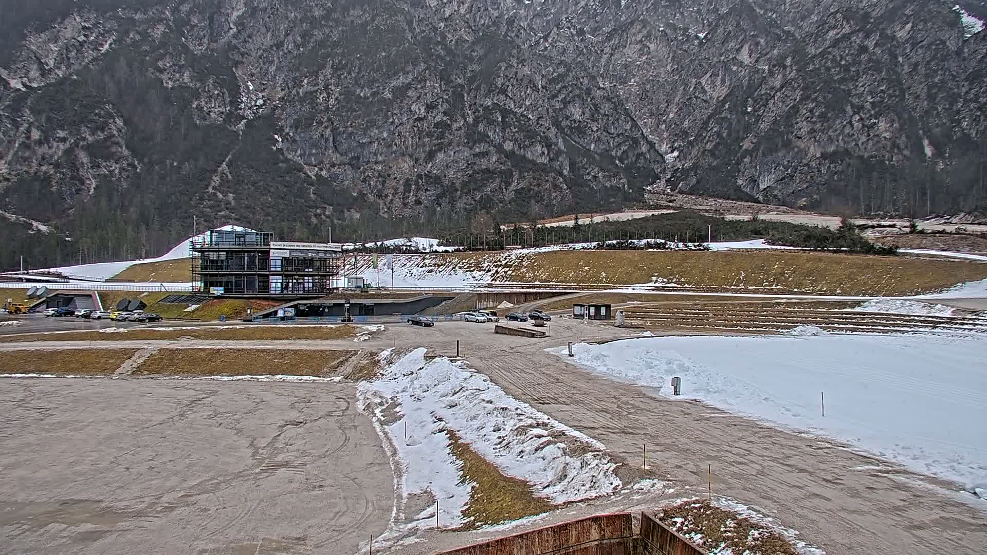 A modern building sits near a snowy, partially dirt track in front of a large, grey mountain under an overcast sky.