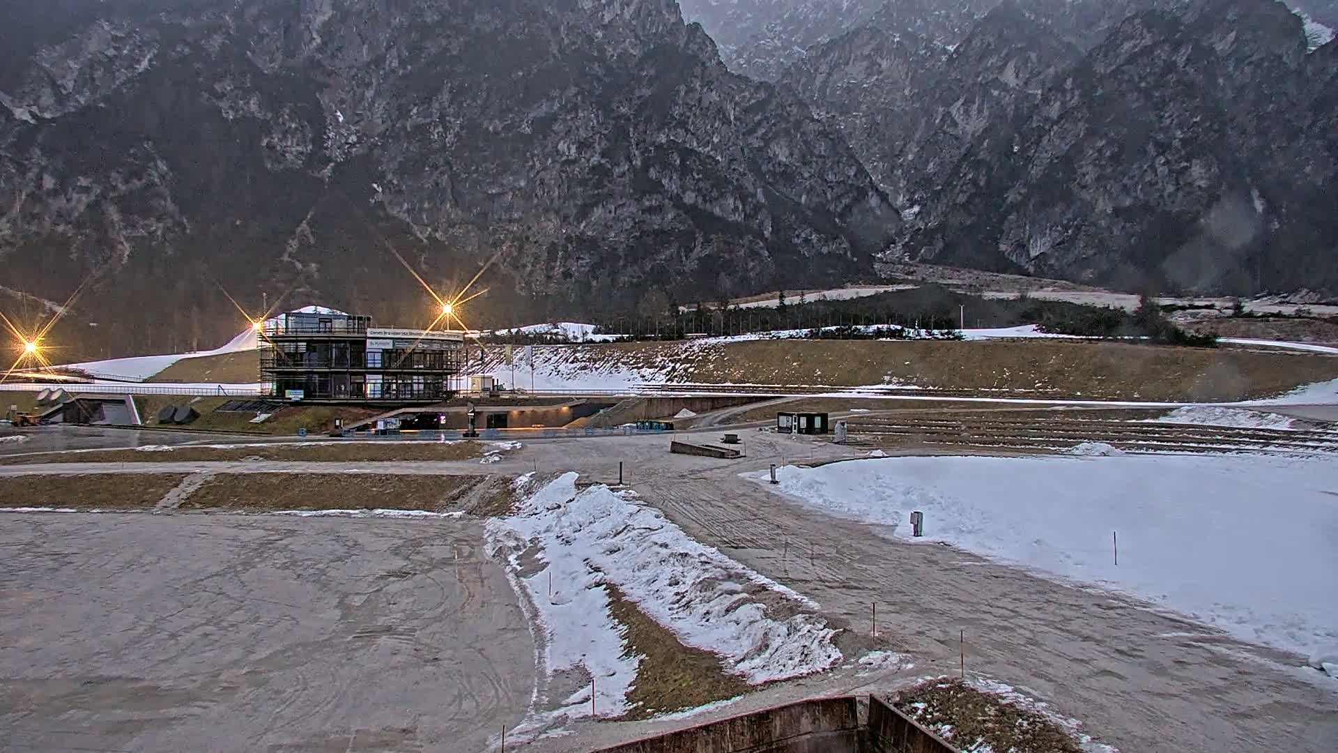 A multi-story building sits in a snowy, mountainous area at dusk, with some areas of exposed ground and several lights illuminating the scene.