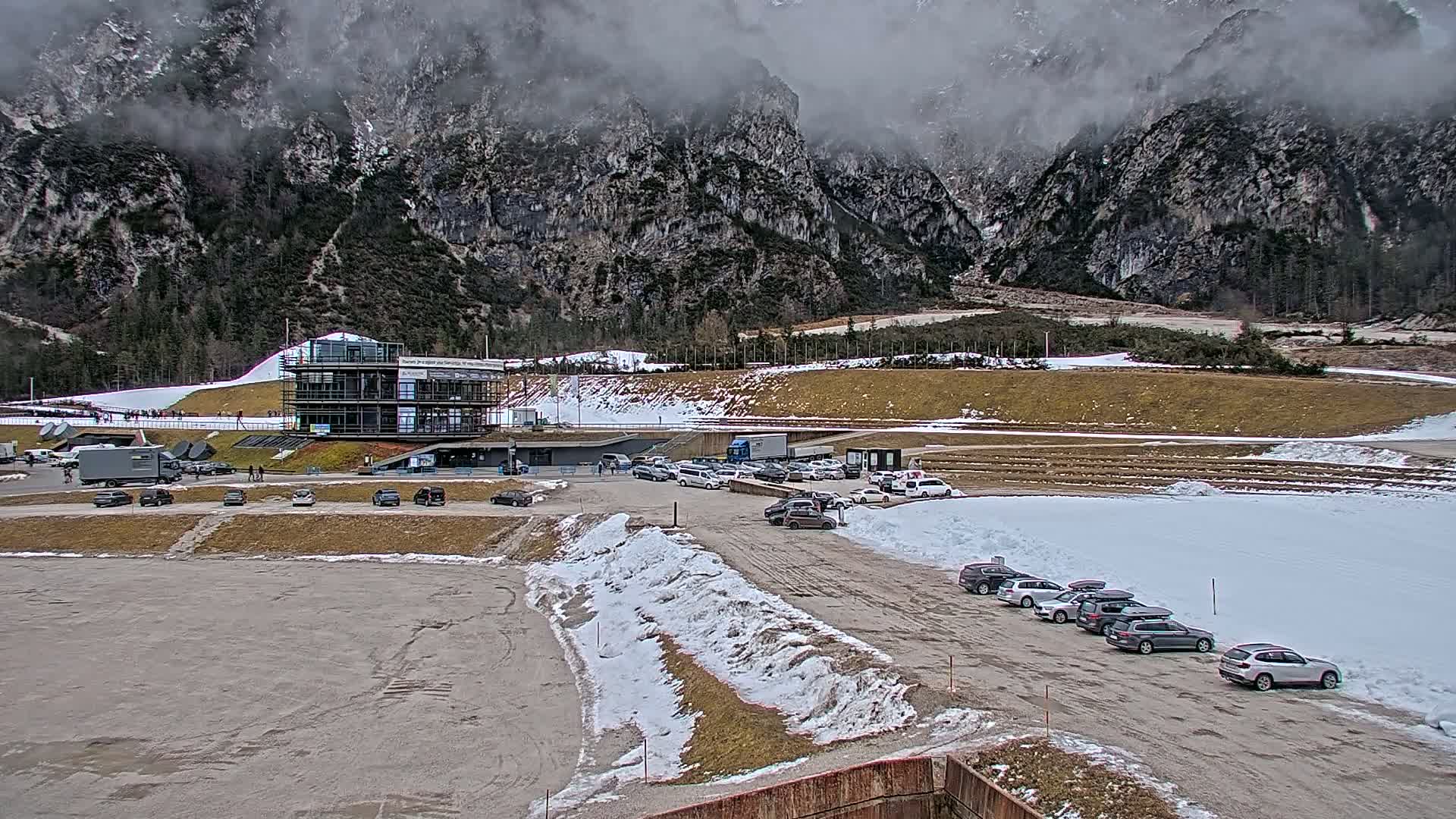 A modern building sits in a parking lot with cars, partially snow-covered ground, and mountains in the background under a cloudy sky.