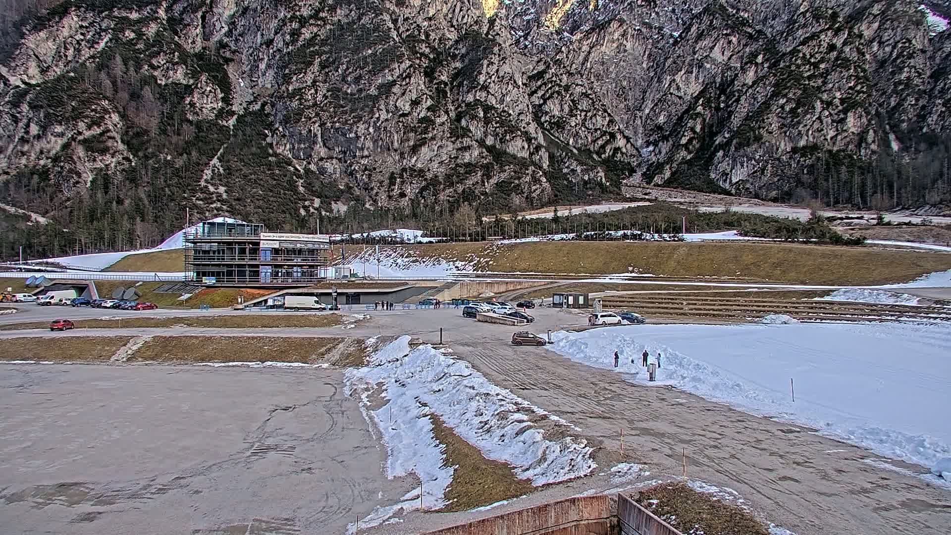 A partially snow-covered parking lot and track sit at the base of a large, rocky mountain on a partly cloudy day.