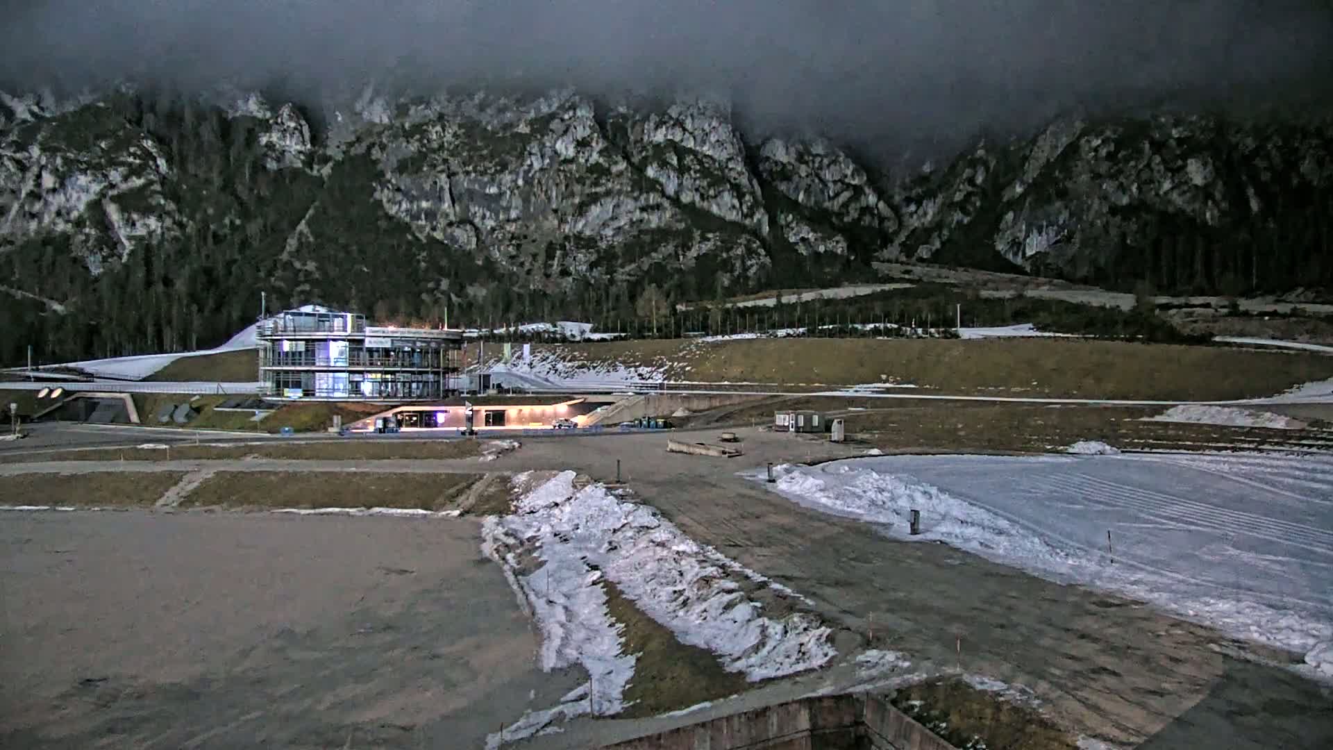 A modern building sits at the base of a large, dark, and foggy mountain range, with patches of snow on the ground surrounding it.