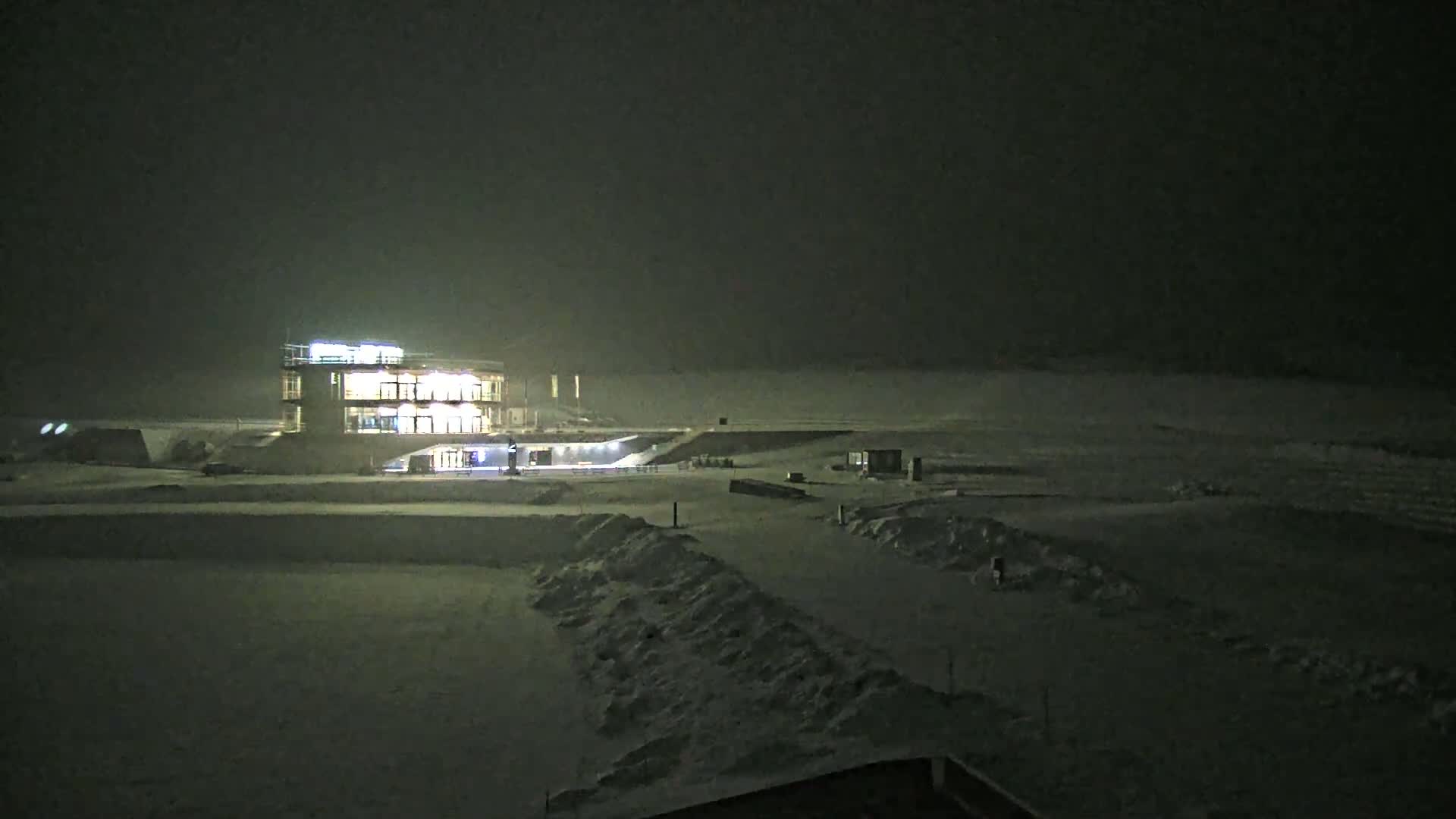 A brightly lit building stands in a snow-covered landscape at night under hazy conditions.