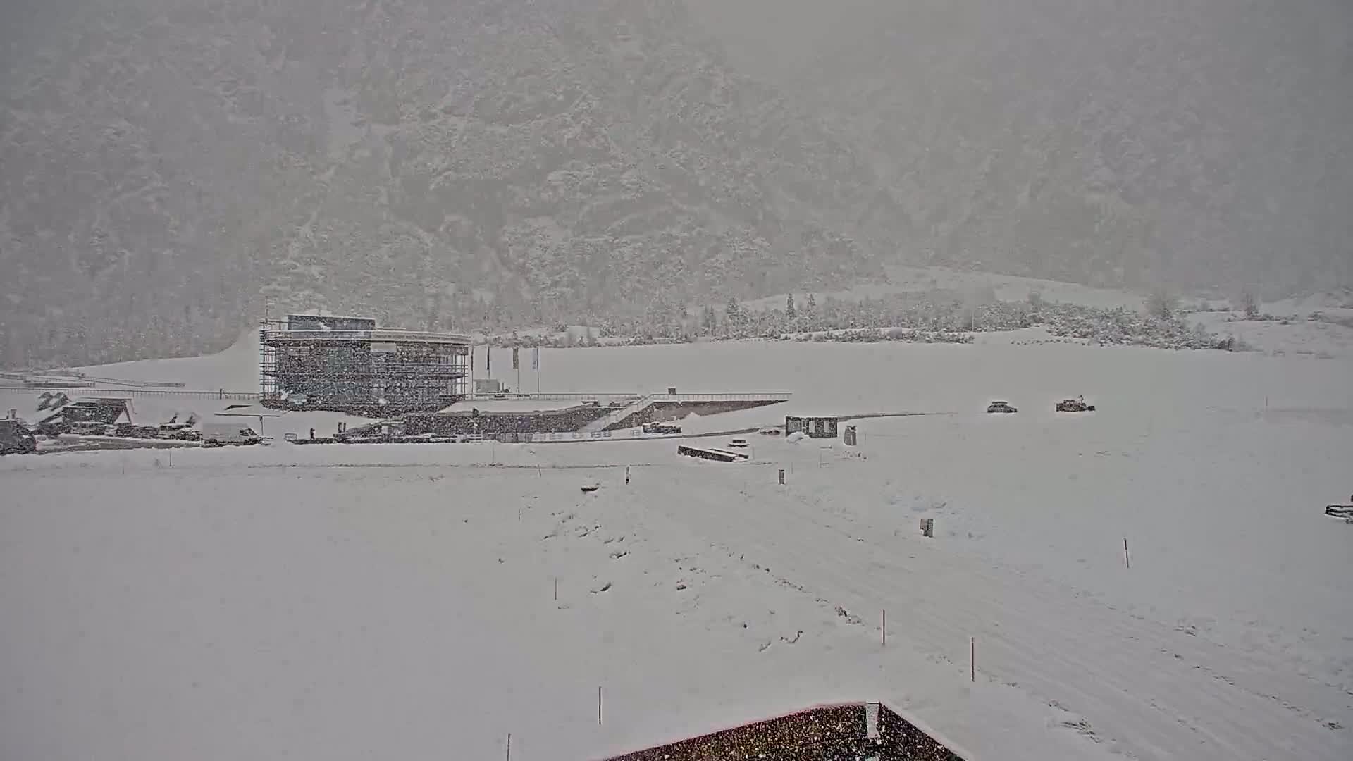 A snow-covered landscape with a partially-constructed building and a few vehicles is seen under heavy snowfall against a mountainous backdrop.