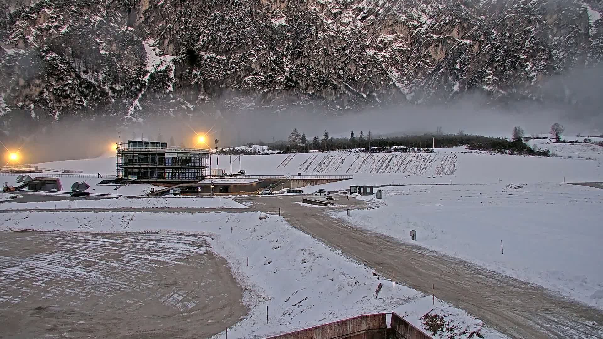 A snowy landscape featuring a modern building near a mountain shrouded in fog, with a few cars parked nearby.