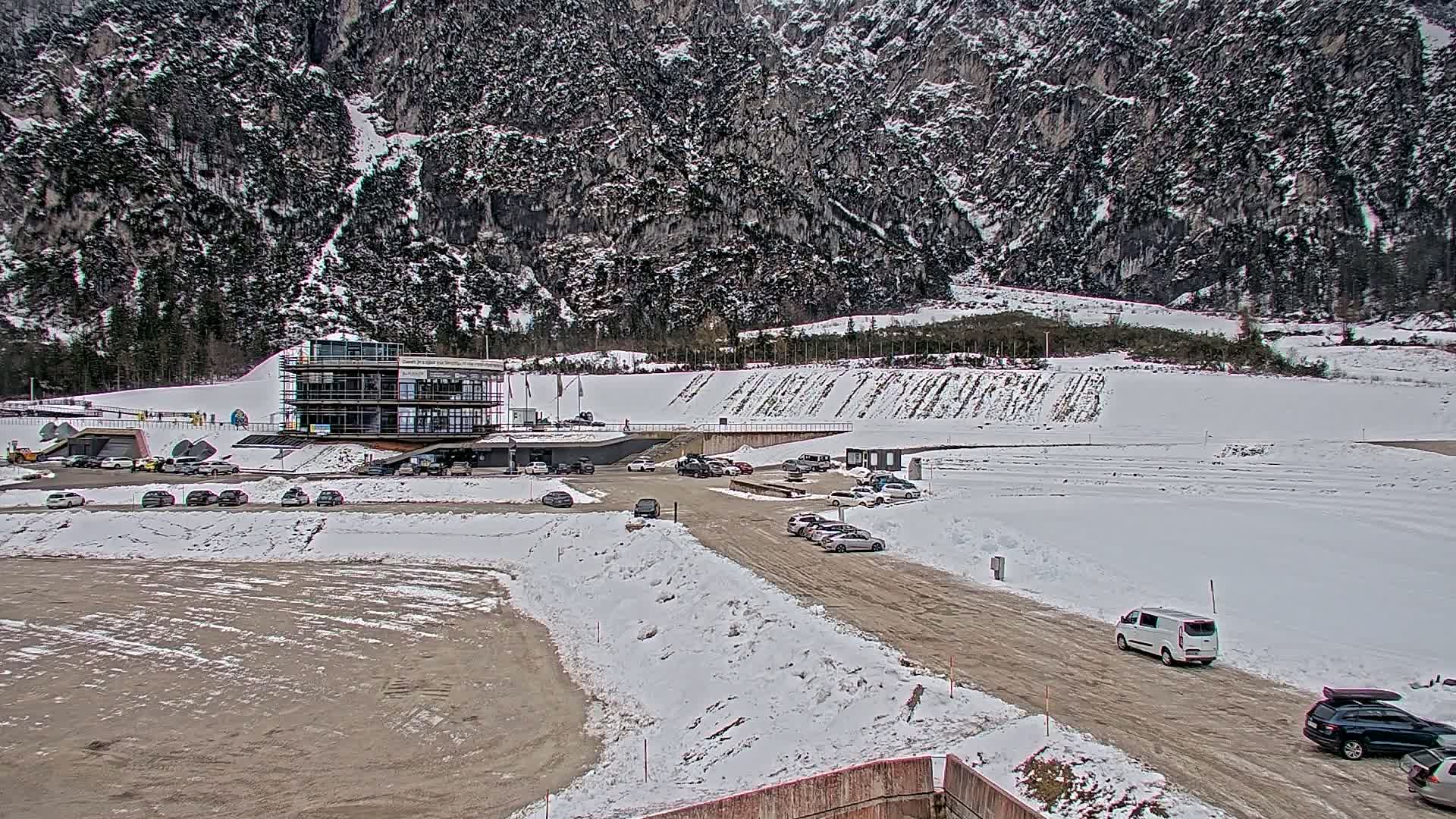 A partially snow-covered parking lot and modern building sit below a large, snow-dusted mountain.