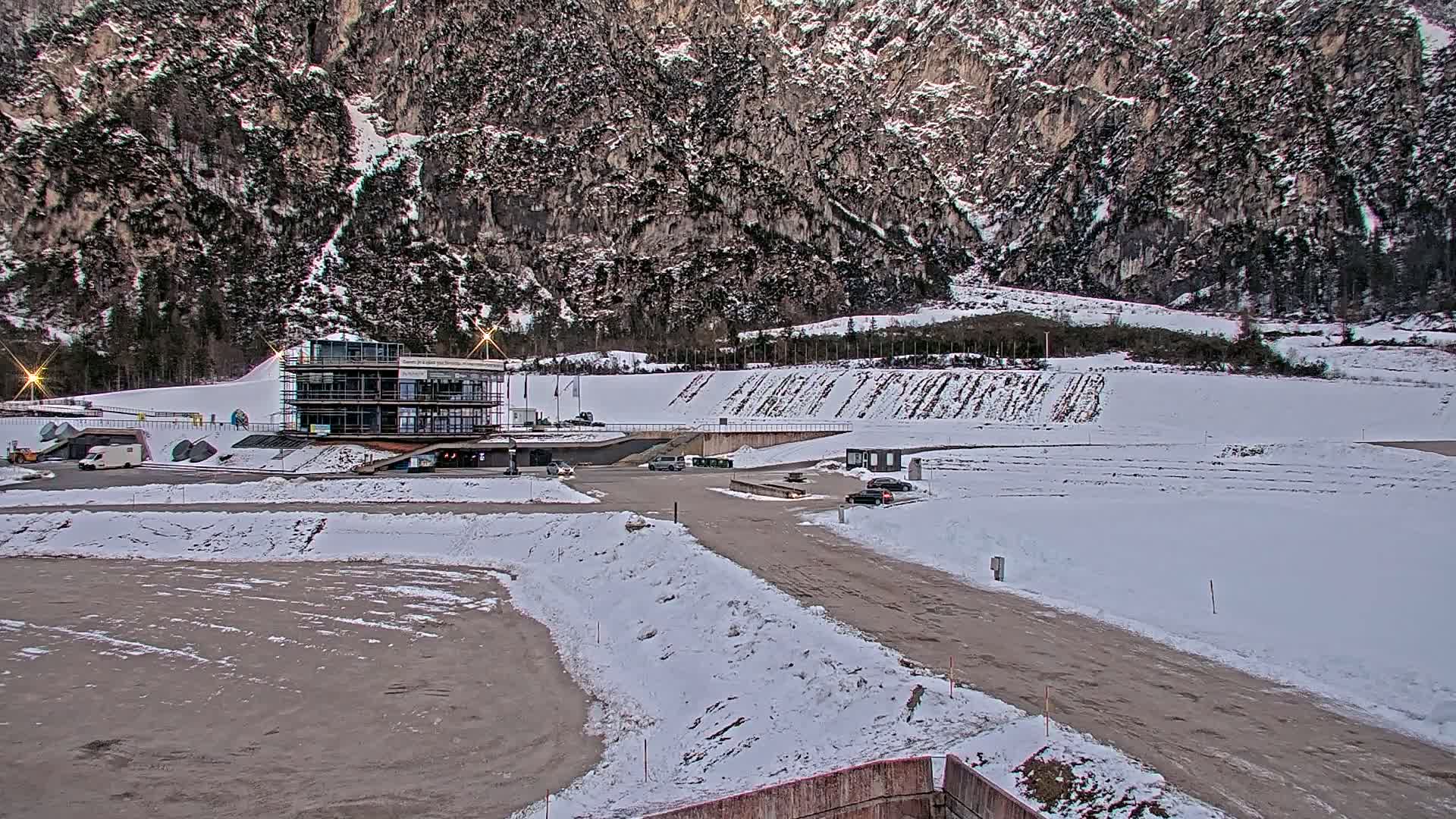 A partially snow-covered parking lot and a modern building sit at the base of a large, snow-dusted mountain range under a cloudy sky.