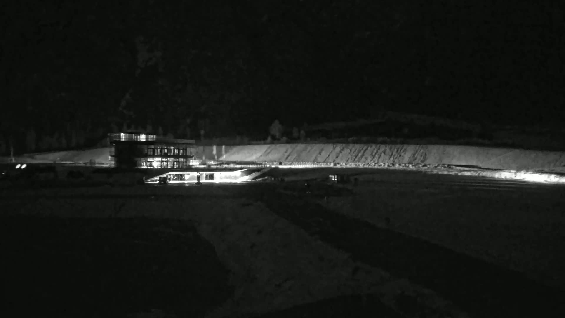 A brightly lit building sits on a snow-covered field at night, with a dark mountain range visible in the background.