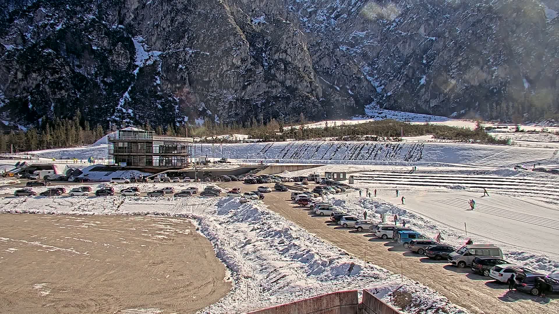 A modern building sits near a snowy parking lot next to a biathlon course set against a backdrop of a large, snow-dusted mountain under a bright, sunny sky.