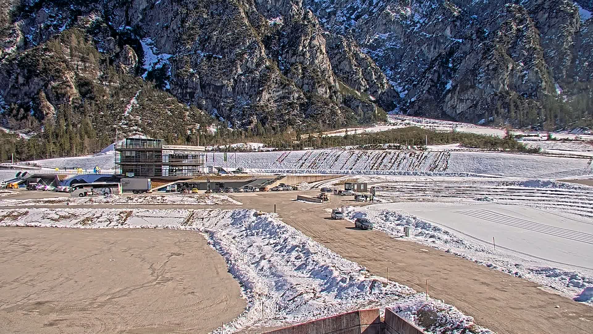 A parking lot with several vehicles is situated in front of a multi-story building, with snowy areas and a mountainous backdrop under a sunny sky.