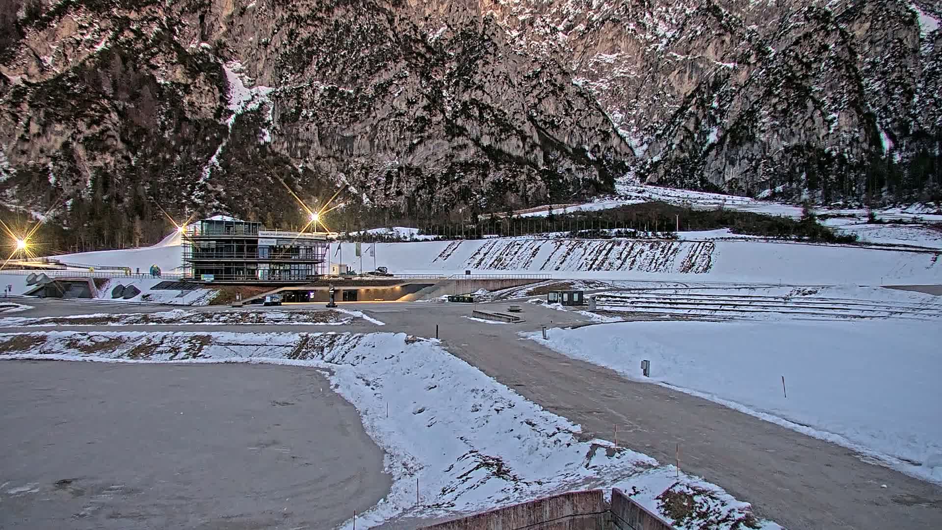 A snow-covered outdoor complex with a multi-story building sits at the foot of a large mountain range under a twilight sky.