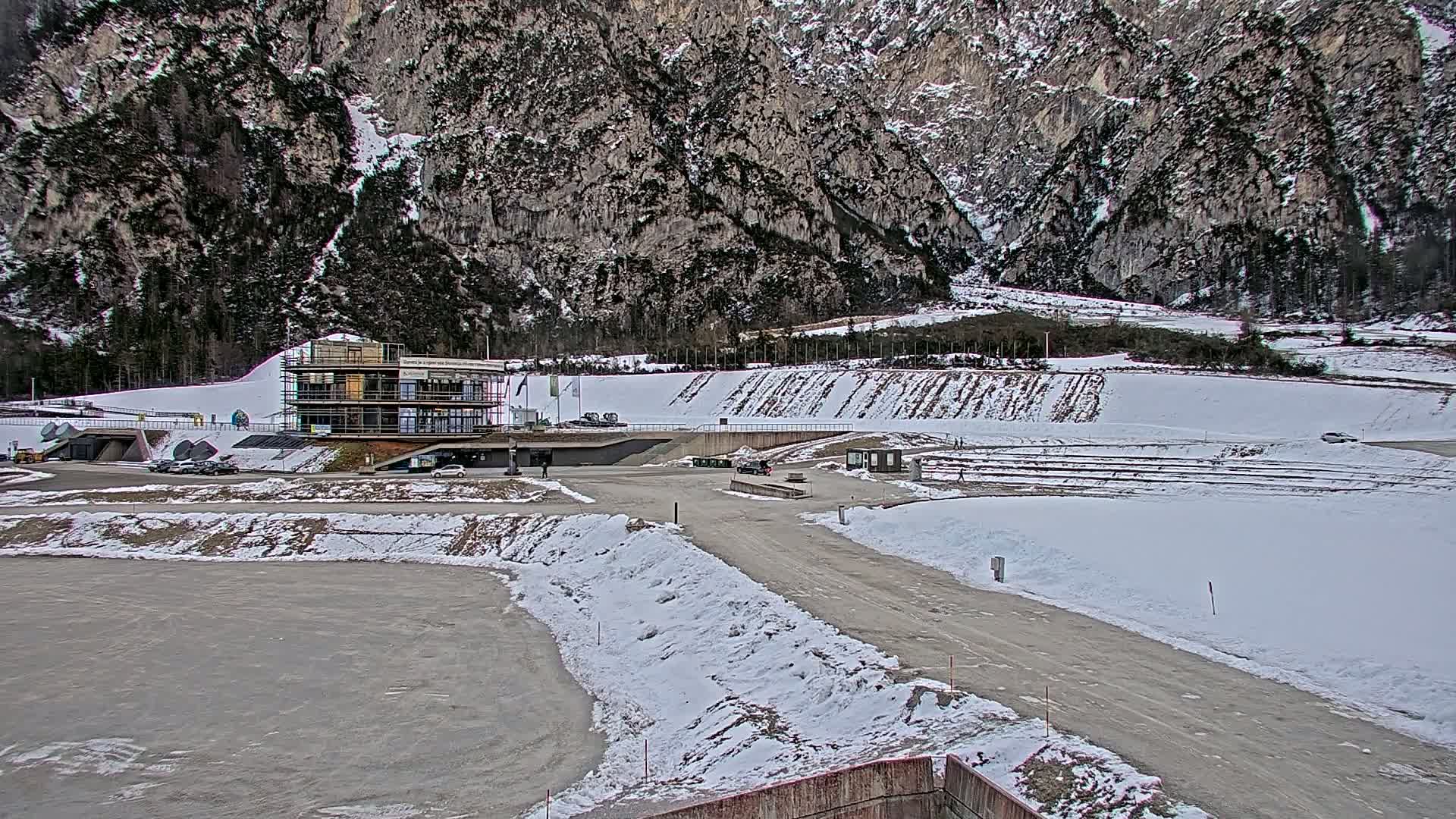 A partially constructed building sits in a snowy parking lot next to a snow-covered athletic field, all set against a backdrop of a large, rocky mountain.