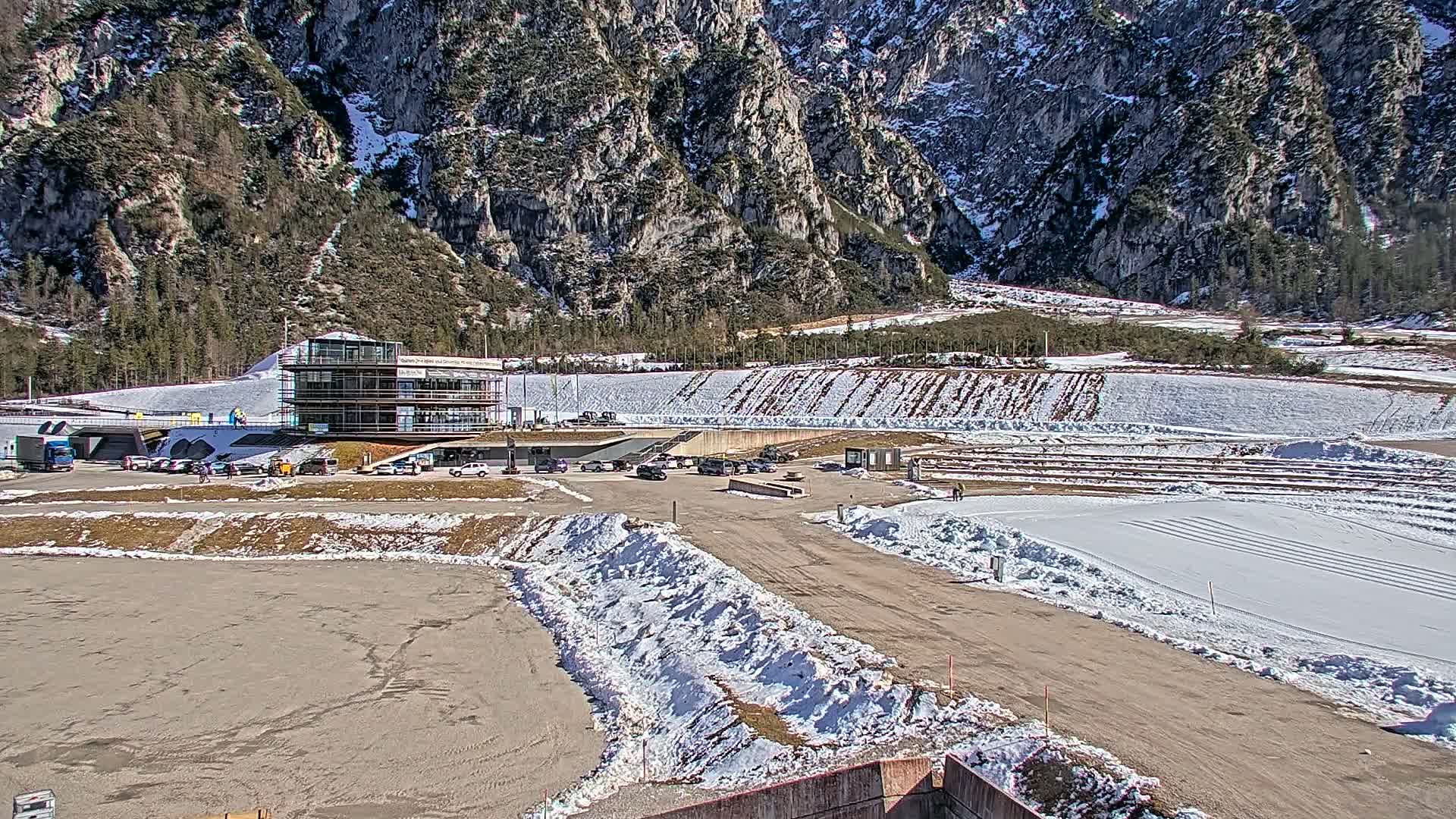 A snow-covered outdoor sporting facility sits at the base of a large, rocky mountain on a sunny day.