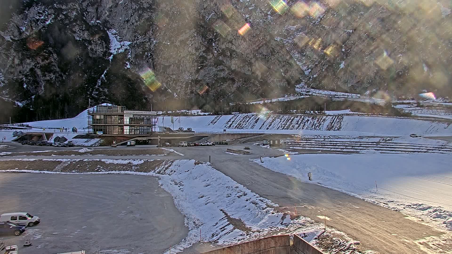 A snow-covered parking lot and roadway are situated in front of a modern building against a backdrop of a mountain under a bright, possibly sunny sky with lens flare.