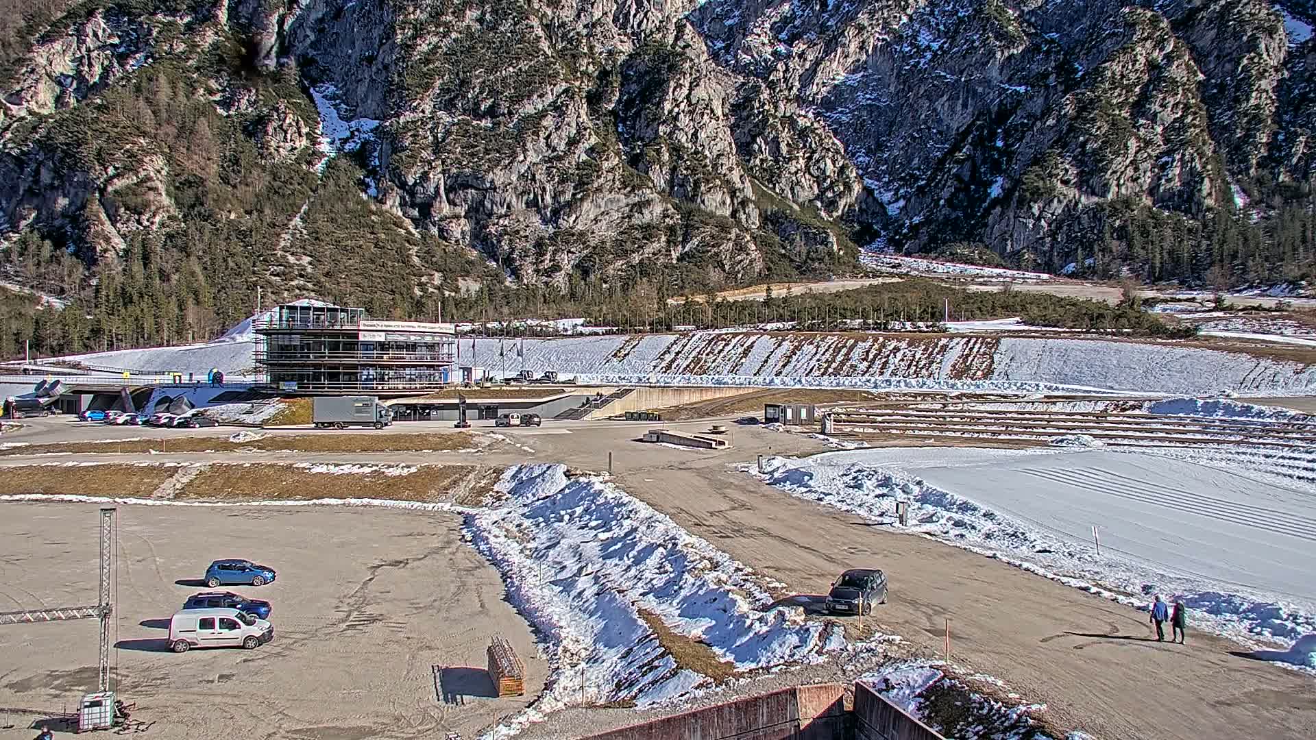 A snowy outdoor complex nestled in a mountainous region, featuring a multi-story building, a parking lot with several cars, and a large, snow-covered track or field.