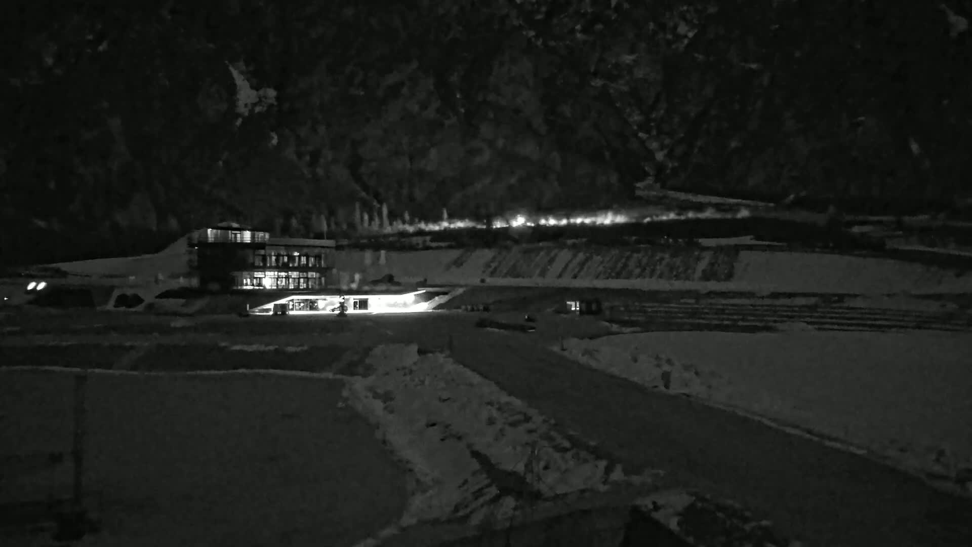 A brightly lit building sits in a snow-covered landscape at night, with a mountain range in the background.