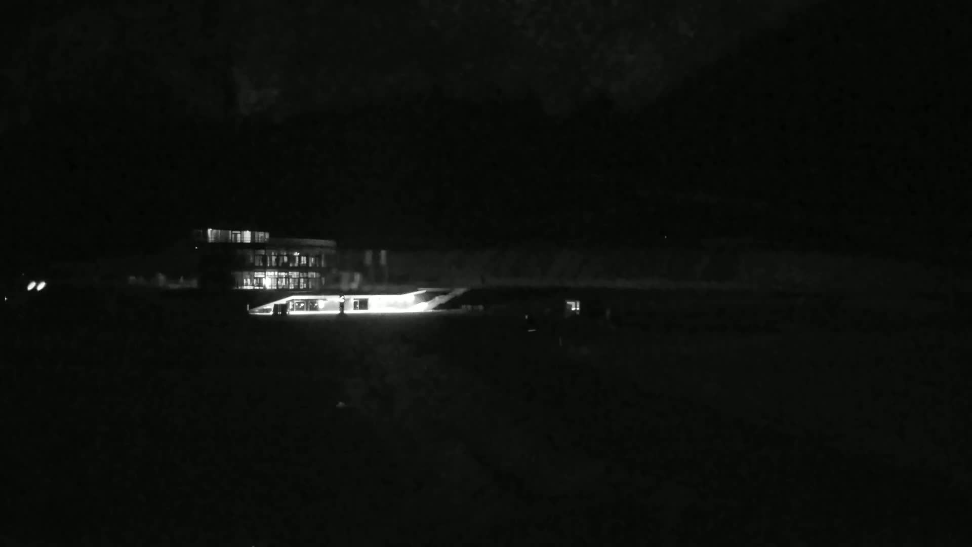 At night, a brightly lit building is visible across a dark, flat expanse of land.