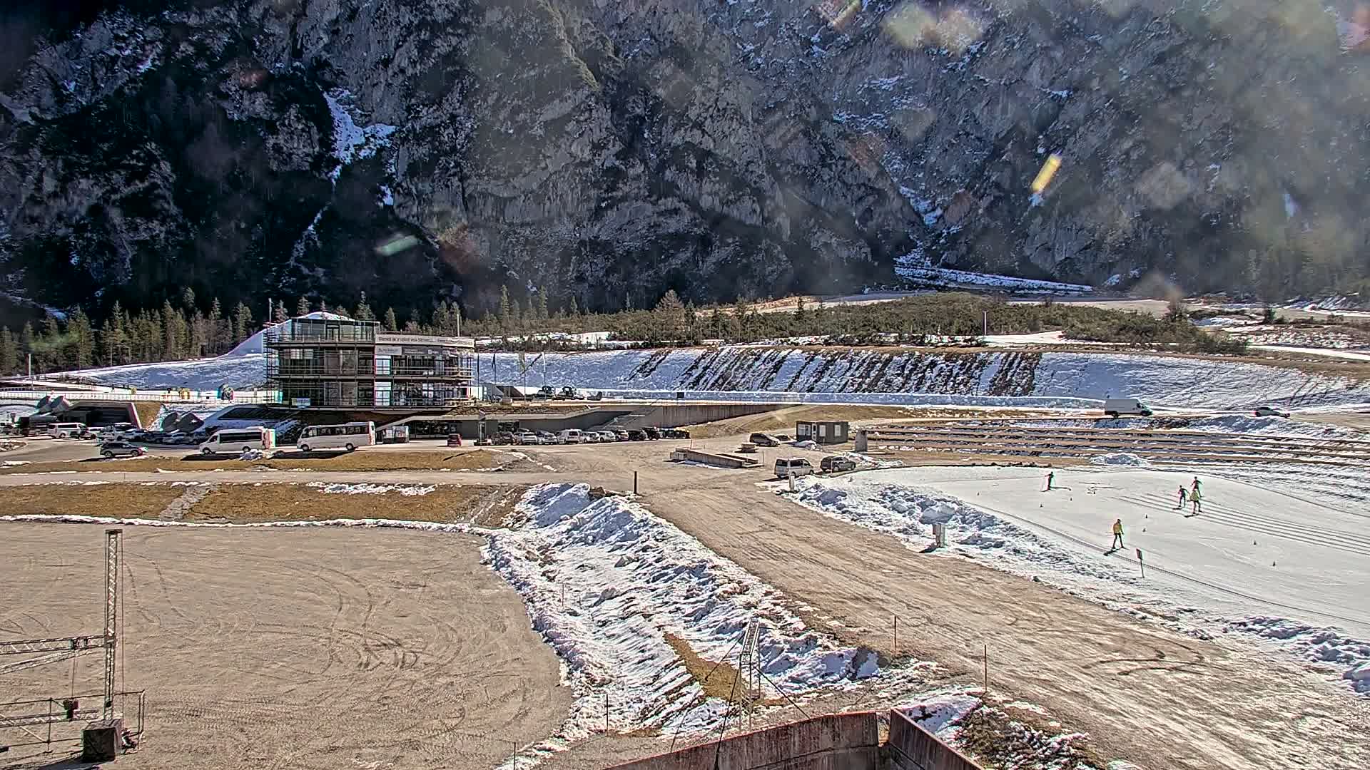A sunny day at a biathlon range nestled in a mountainous valley, showing a building, parking lot, and several people cross-country skiing on a snow-covered course.
