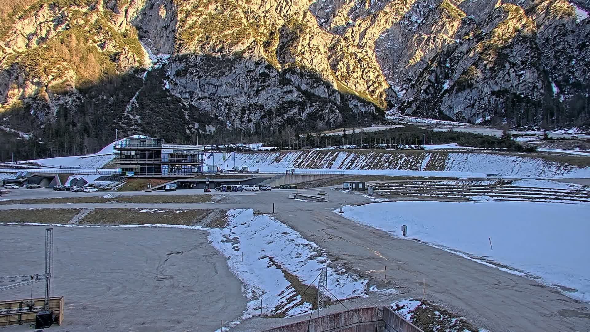 A partially snow-covered parking lot and athletic facility are situated in a valley, backed by a steep, rocky mountainside under a partly sunny sky.