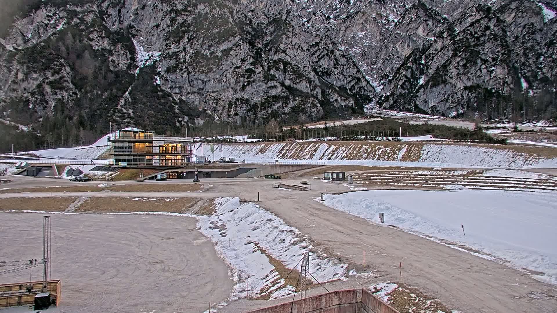 A partially snow-covered outdoor sports facility, featuring a multi-story building, is situated at the base of a large, rocky mountain.