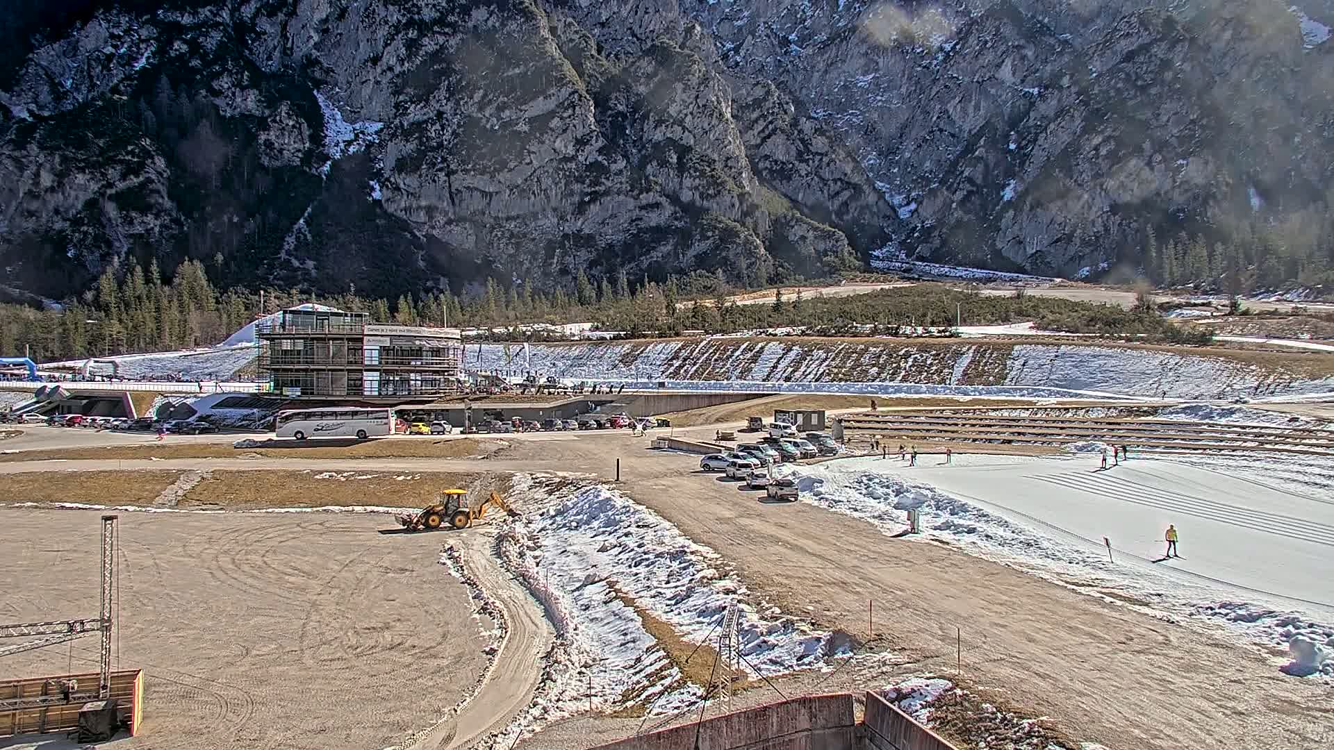 A sunny day reveals a snowy outdoor sporting venue nestled in a valley between large, rocky mountains, with a building, vehicles, and a few people visible.