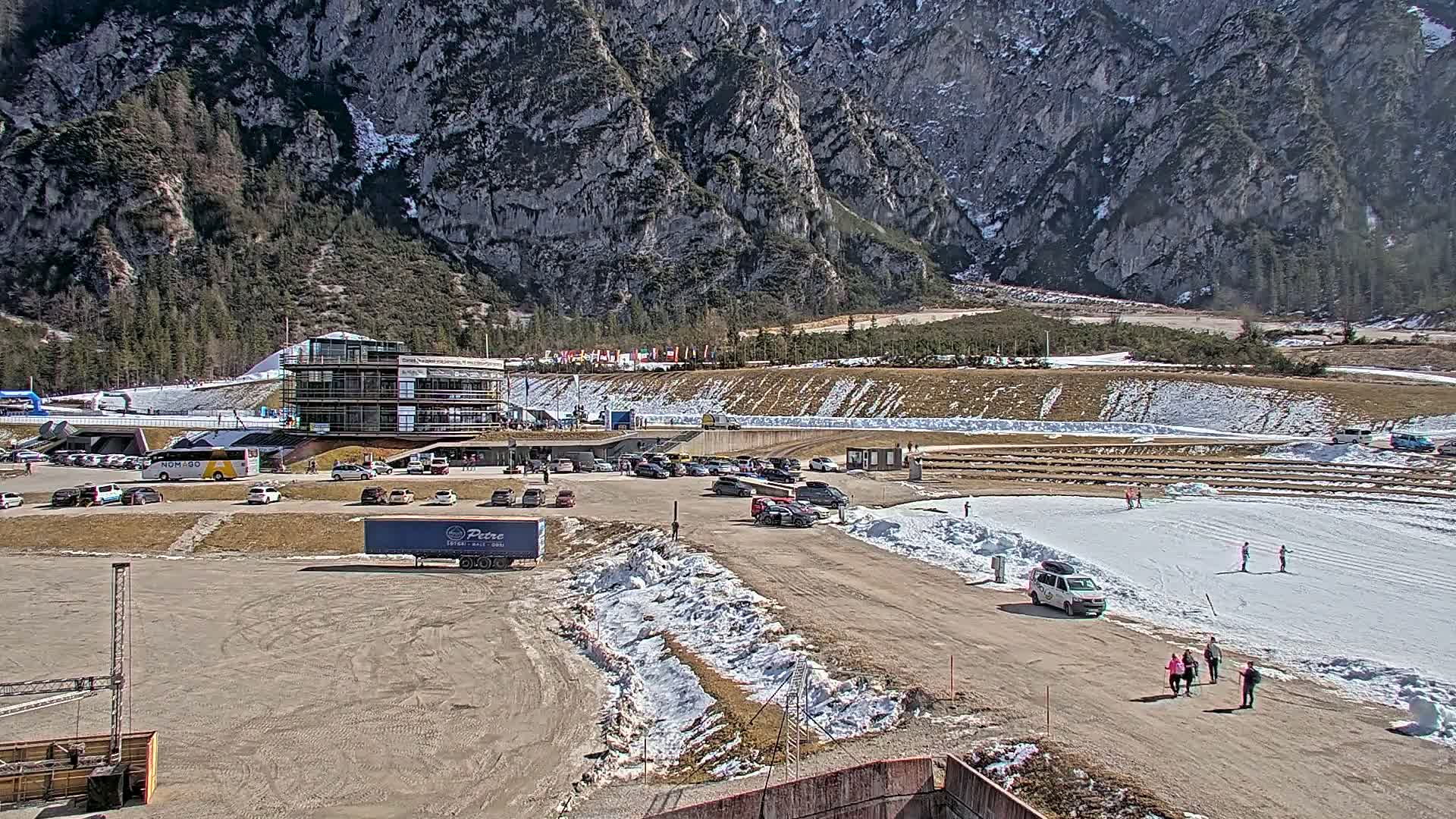 A parking lot with cars and a large building sits in front of a snowy, mountainous landscape on a sunny day.