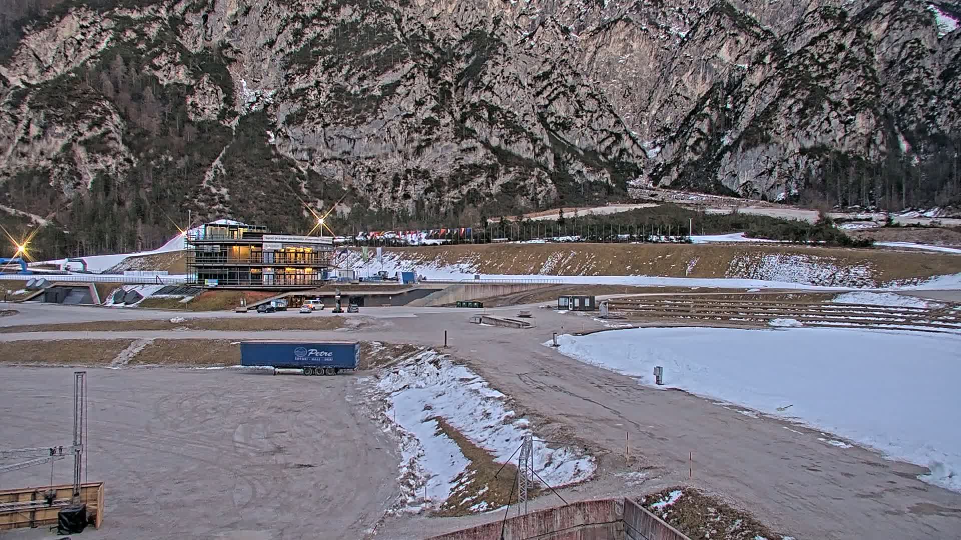 A large, mostly empty parking lot with patches of snow sits below a multi-story building near a snowy ski jump in front of a large, rocky mountain under a mostly clear sky.