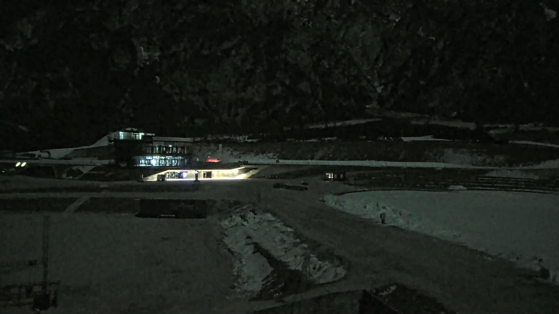 A snow-covered nighttime scene shows a lit-up building near a dark mountain range.
