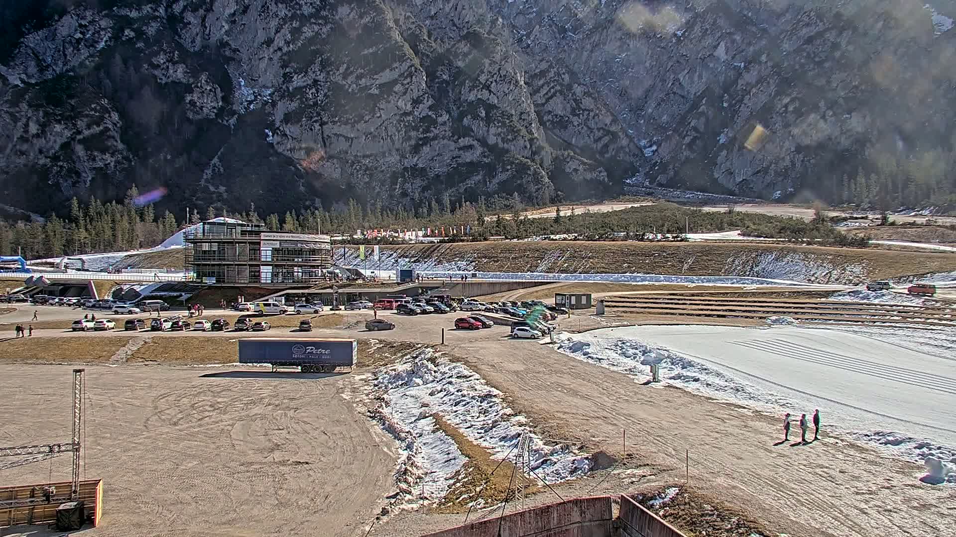 A parking lot with cars and a multi-story building is situated at the base of a large, rocky mountain on a sunny day with patches of snow on the ground.