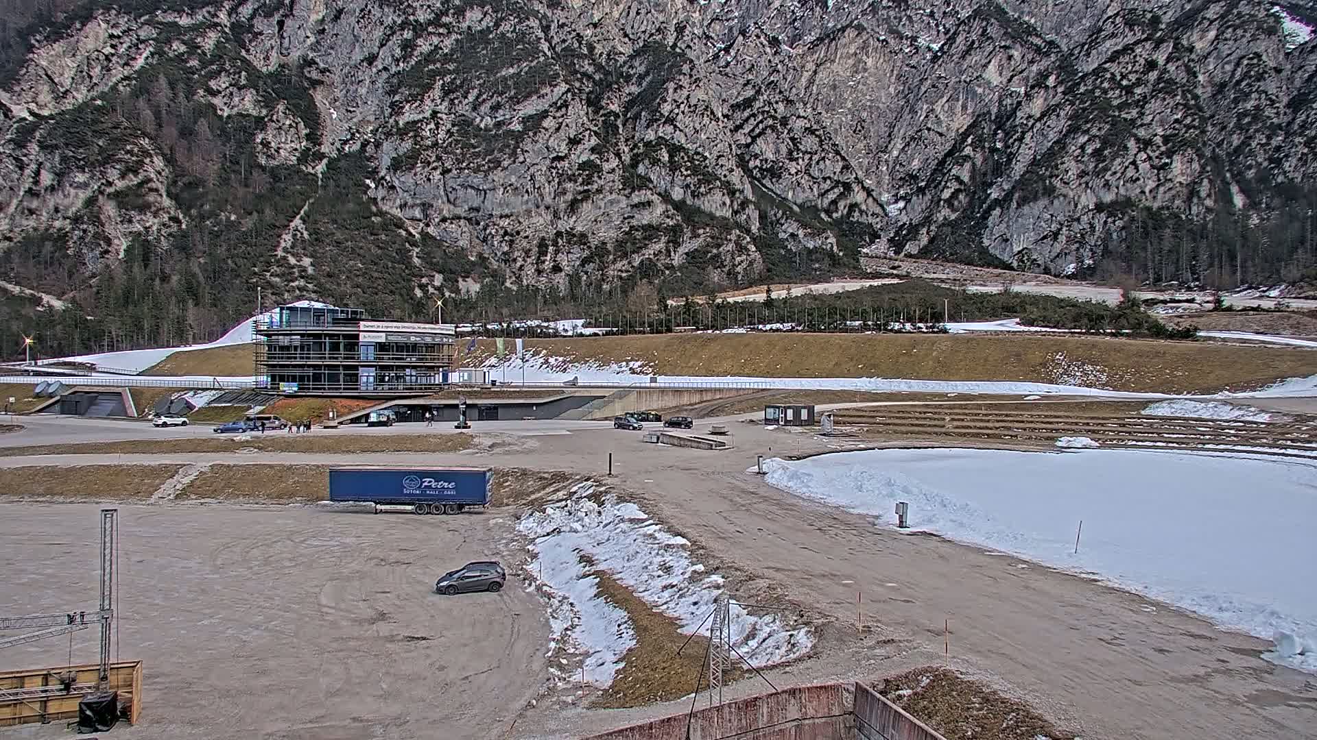 A large, multi-story building sits in a parking lot with patches of snow and dirt, adjacent to a snowy, sloping track against a backdrop of a large, rocky mountain under a cloudy sky.