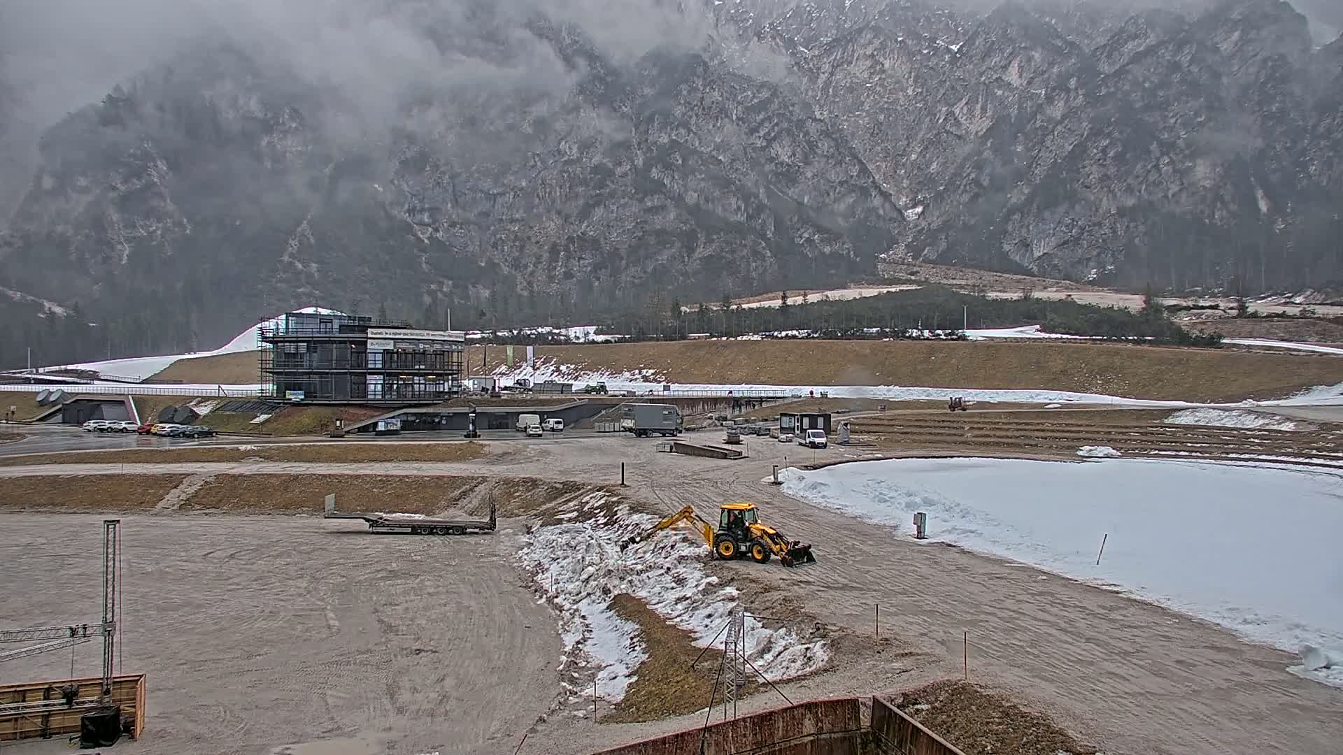A cloudy day reveals a construction site with a yellow excavator working near a snow-covered area, in front of a multi-story building and a large mountain range in the background.