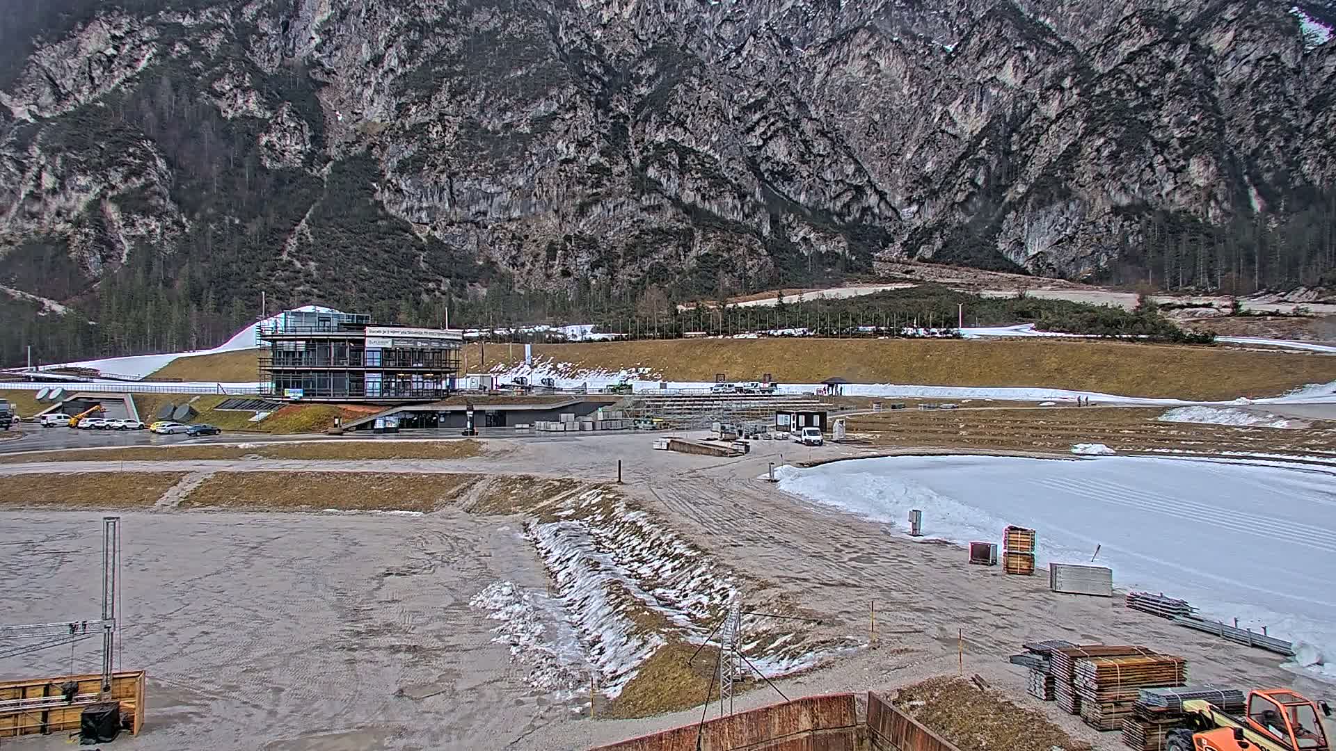 A partially snow-covered outdoor facility with a multi-story building, surrounded by a mountain backdrop under overcast skies.