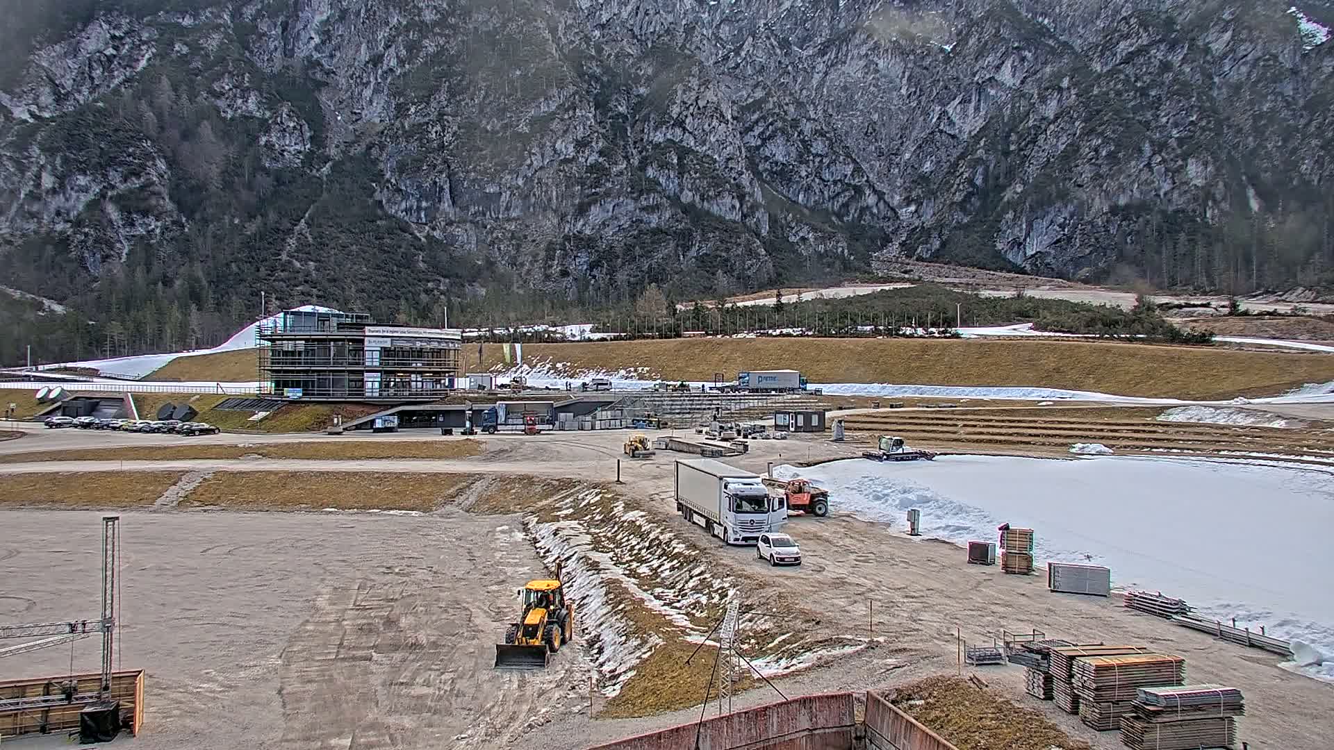 A large construction site with several trucks, pieces of heavy equipment, and stacks of lumber is situated in front of a multi-story building near a snow-covered mountain under an overcast sky.