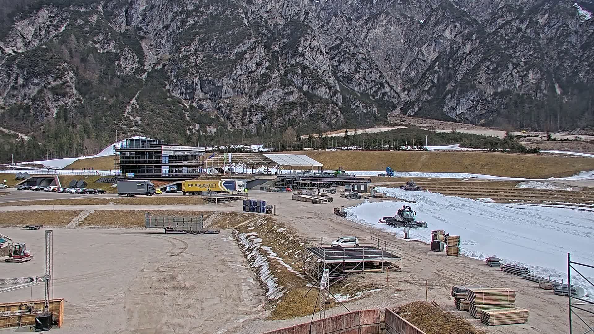 A partially snow-covered outdoor area with construction equipment and vehicles near a multi-story building, set against a backdrop of a large mountain range under a cloudy sky.