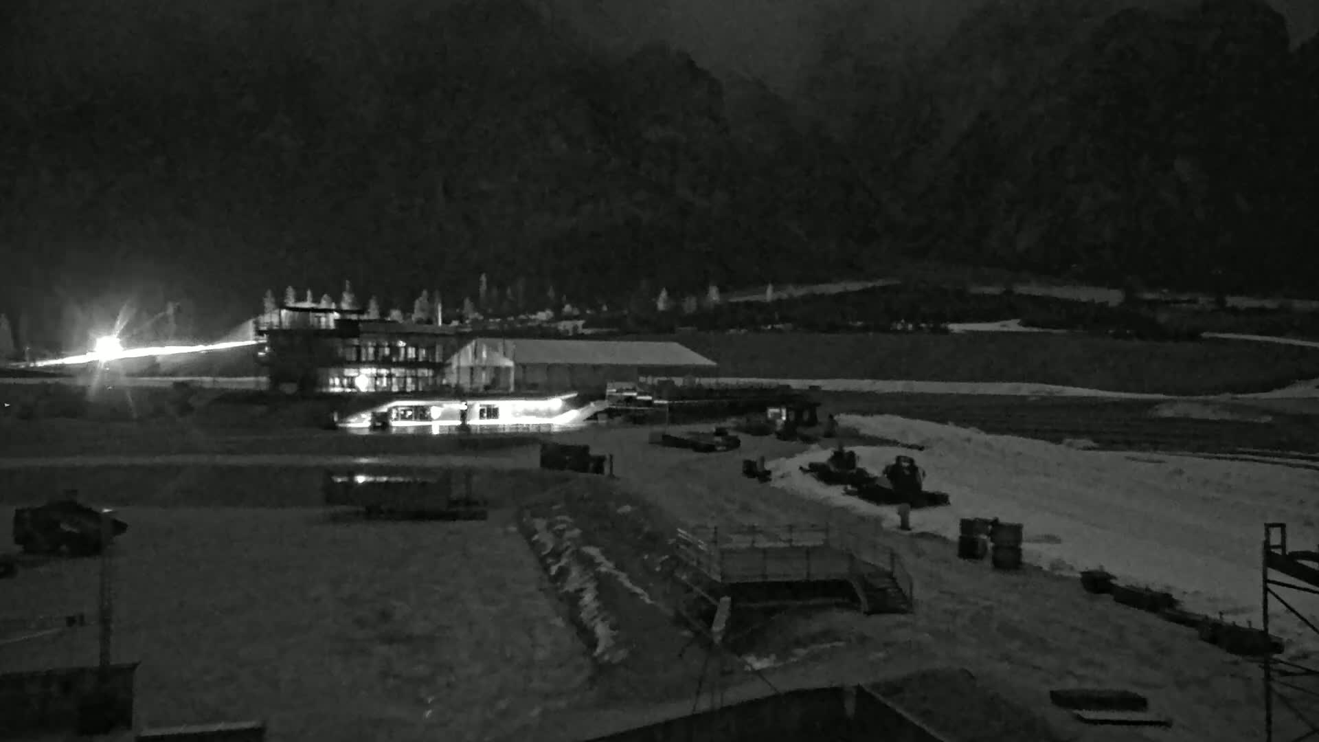 A nighttime, snow-covered landscape features a lit-up building complex and various pieces of machinery near a mountain range under a dark sky.