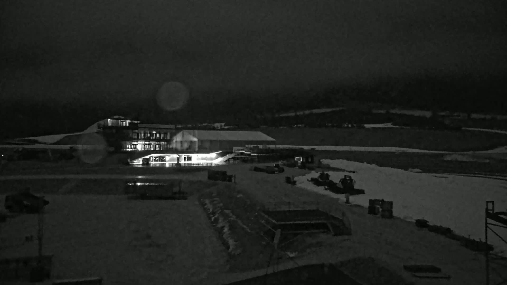 A brightly lit building complex sits in a snow-covered landscape at night under a dark, cloudy sky.