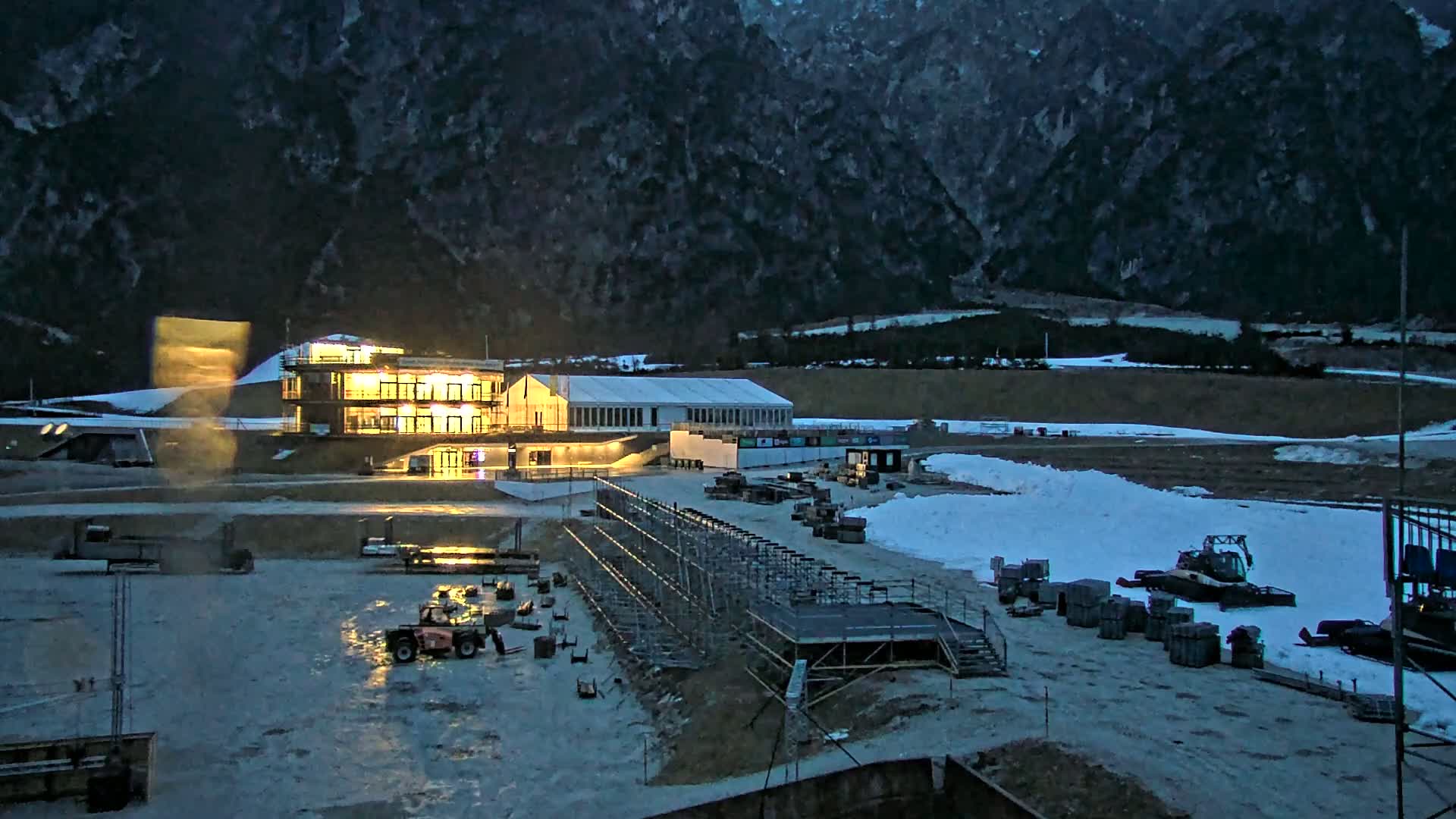 A brightly lit building and a large white tent sit in a snowy outdoor area at dusk, against a backdrop of dark mountains.
