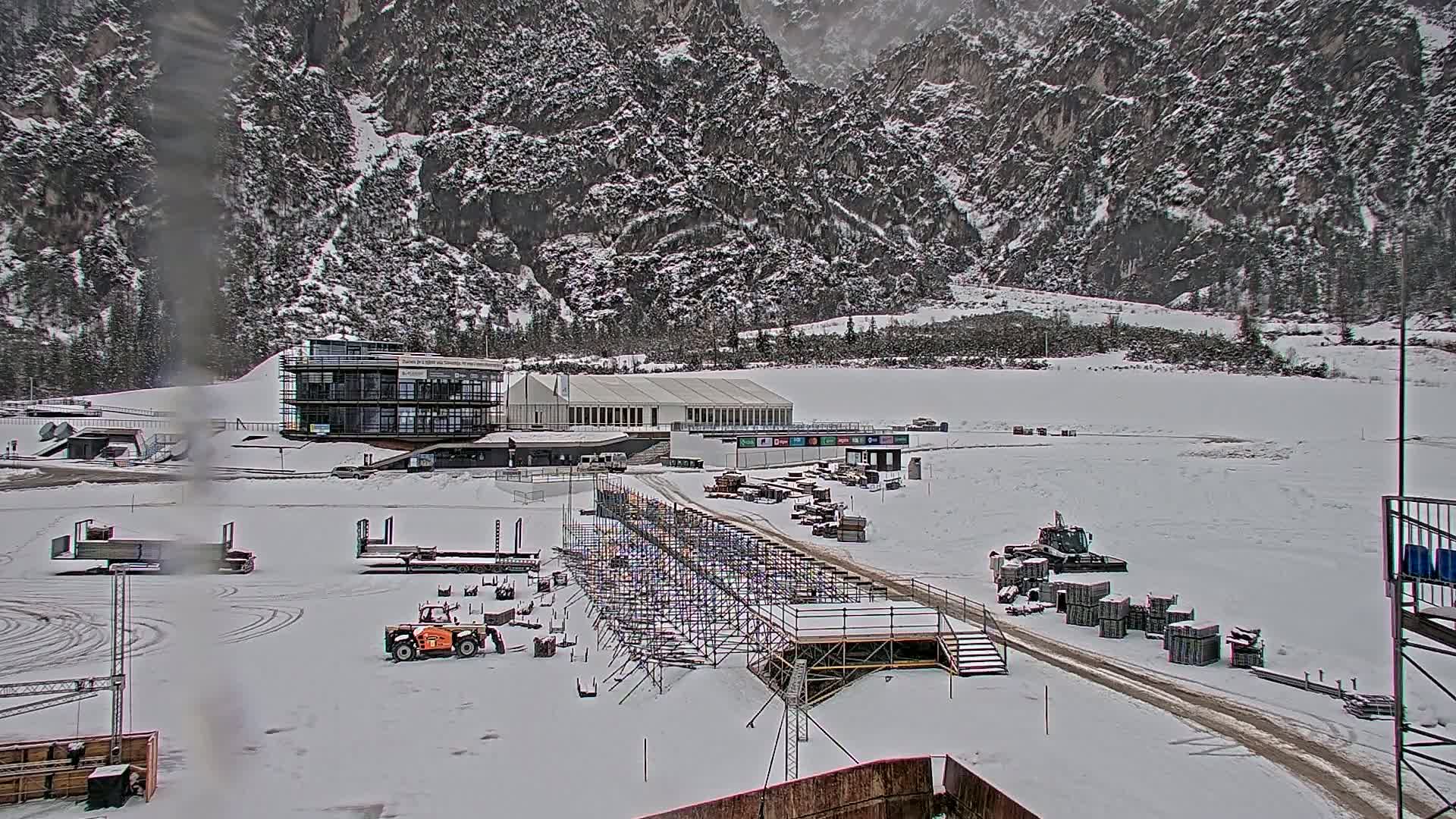 A snowy landscape shows a large metal scaffolding structure, several pieces of construction equipment, and buildings near snow-covered mountains.