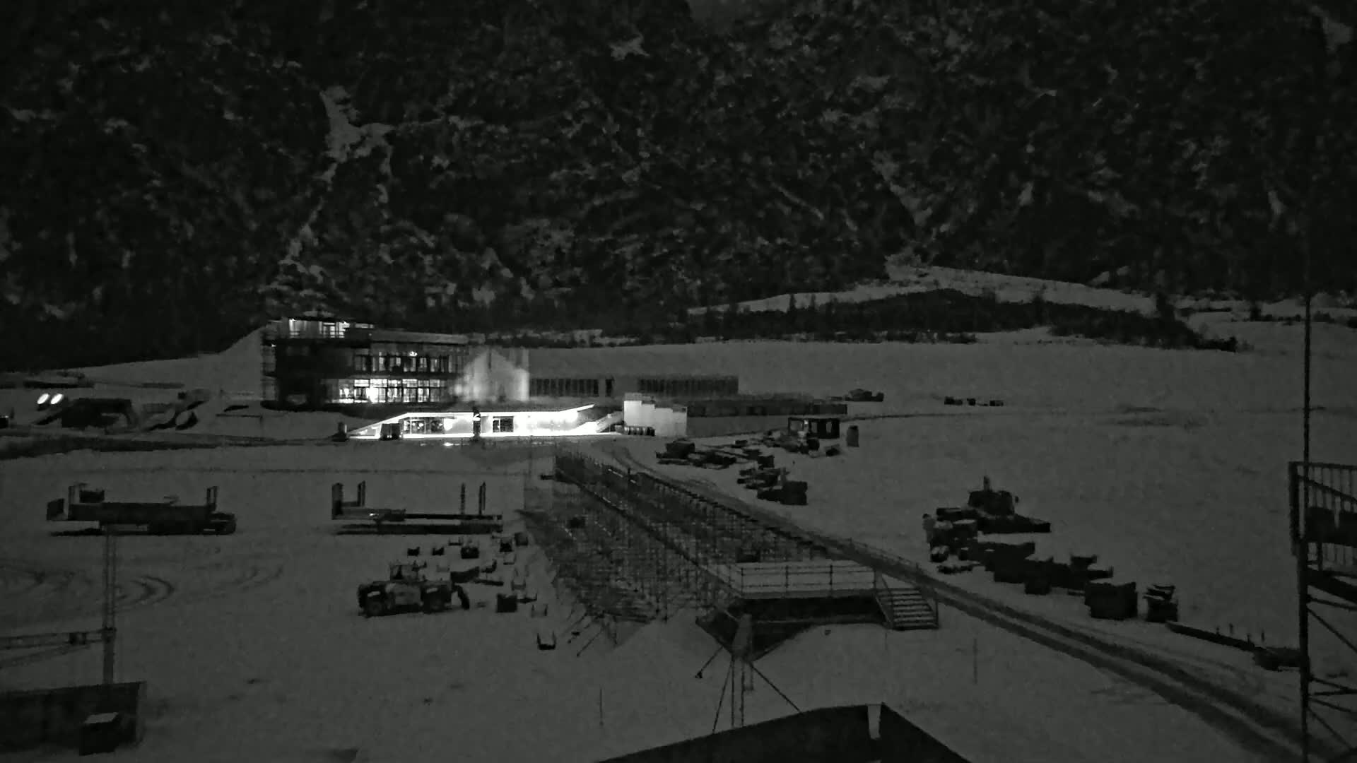 A snow-covered nighttime scene shows a lit building and various equipment near a mountain under a dark sky.