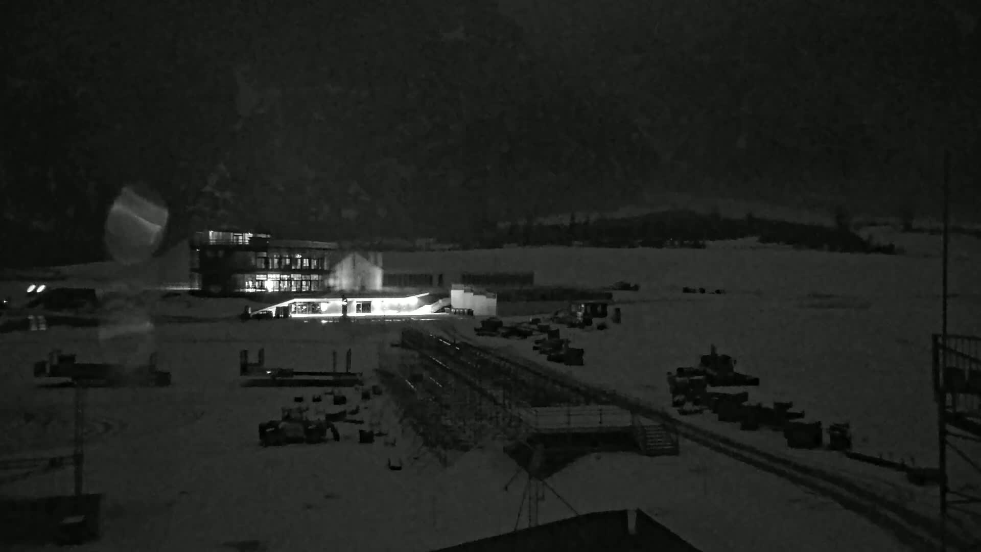 A nighttime, snow-covered scene shows a lit building and various pieces of machinery scattered across a large, flat area.