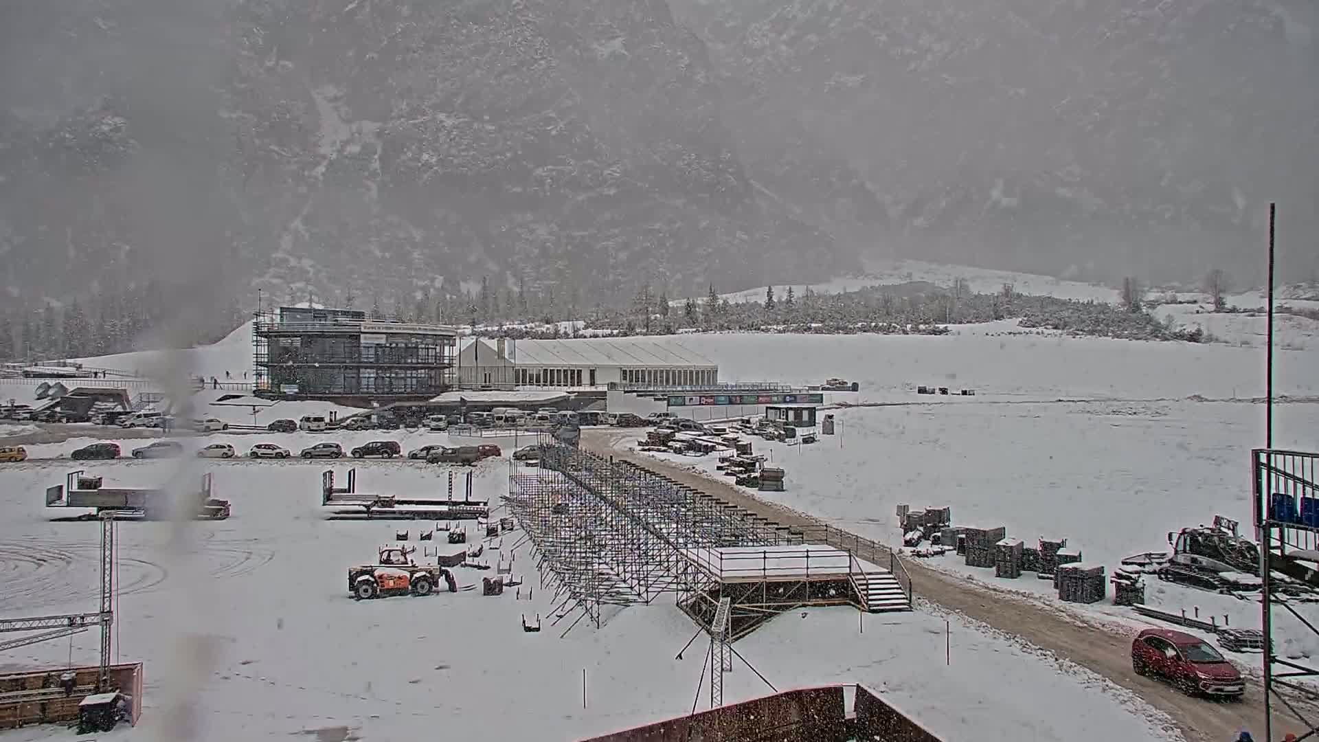 A snow-covered outdoor scene shows a large building complex, vehicles, and construction equipment near a mountain range during a snowfall.